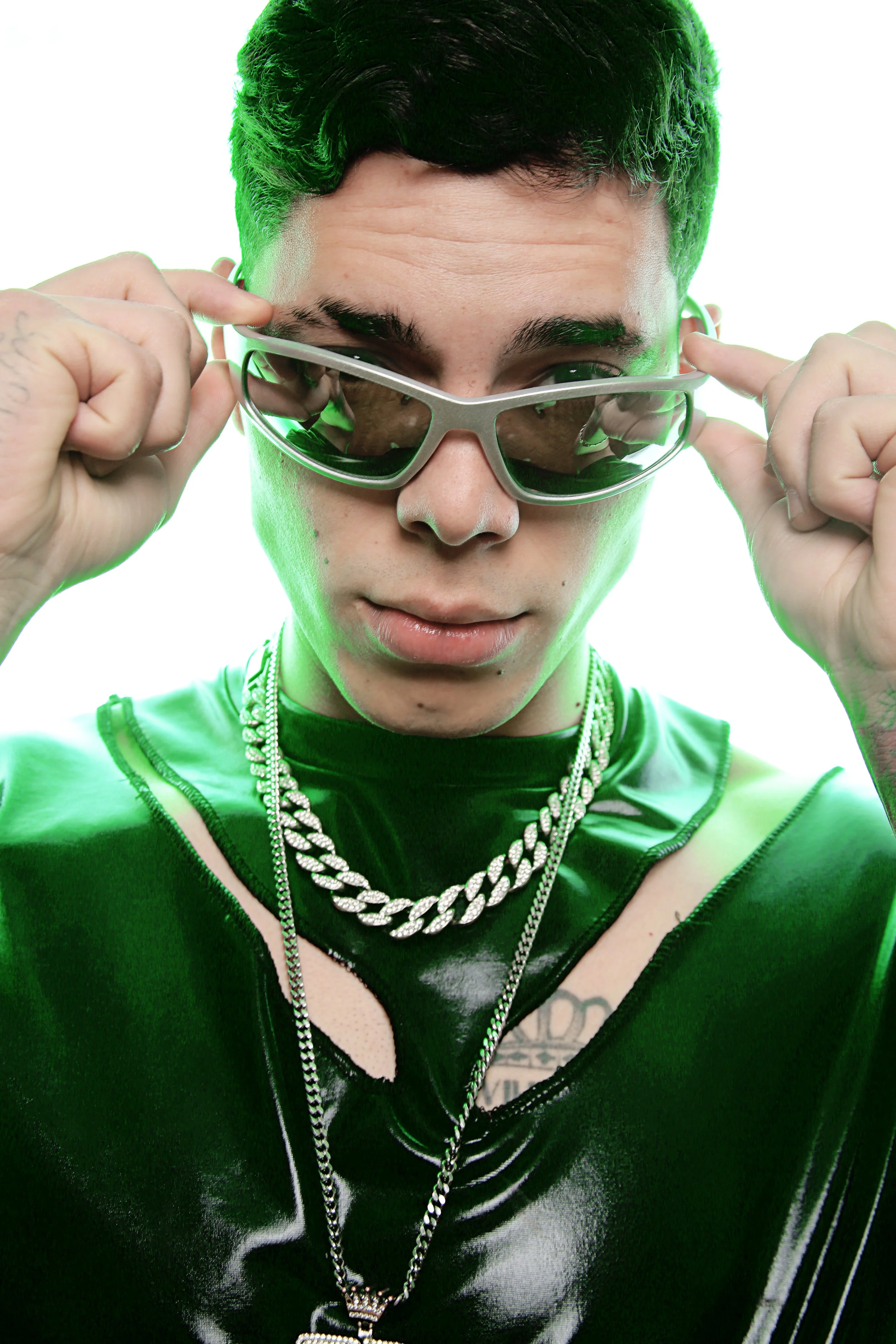 Close-up of a person with short black hair, wearing tinted sunglasses, a shiny black vest, layered silver chains, and a tattoo on their chest with visible text.