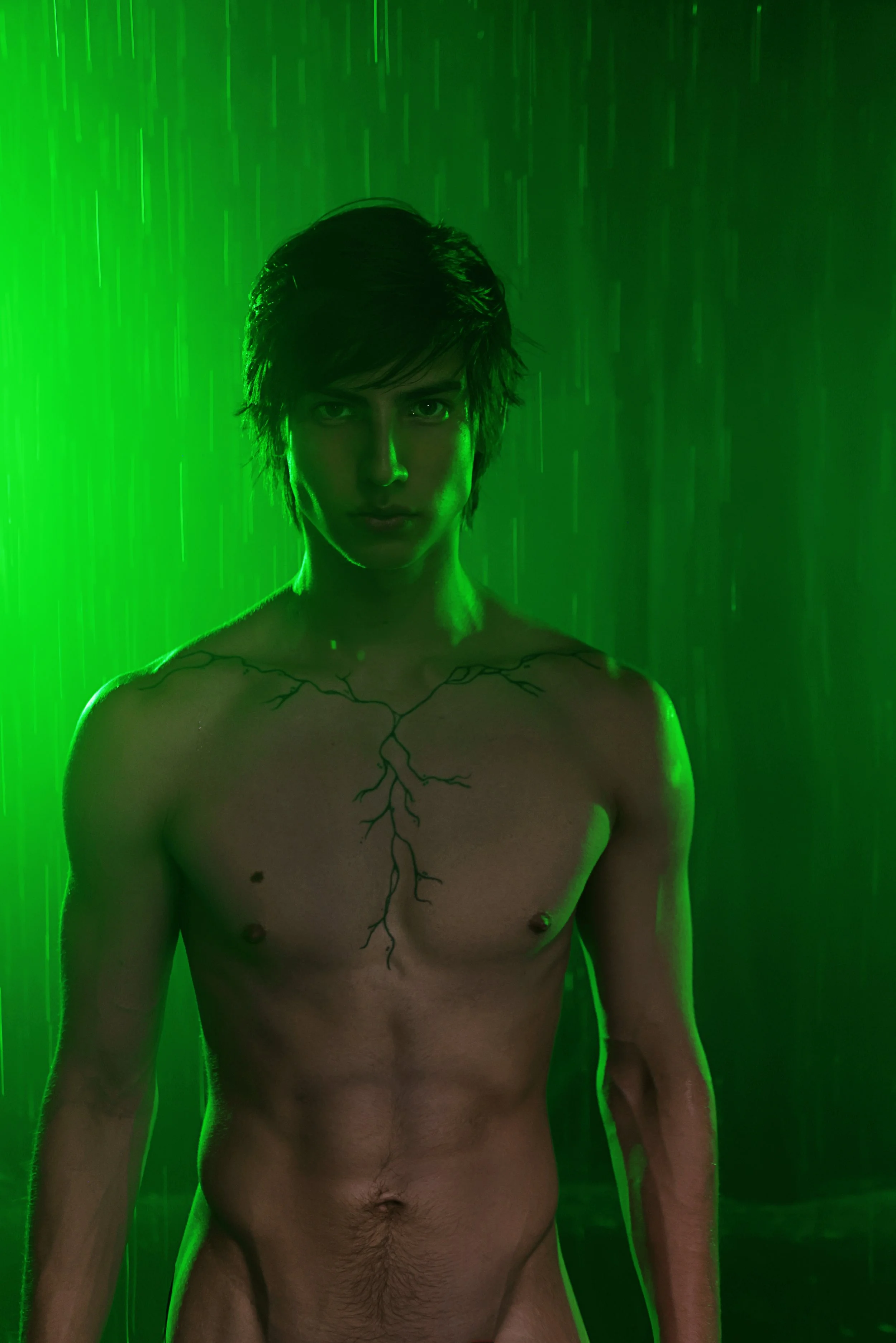 A shirtless man with black hair and a vine tattoo on his chest standing in front of a green background with rain falling.