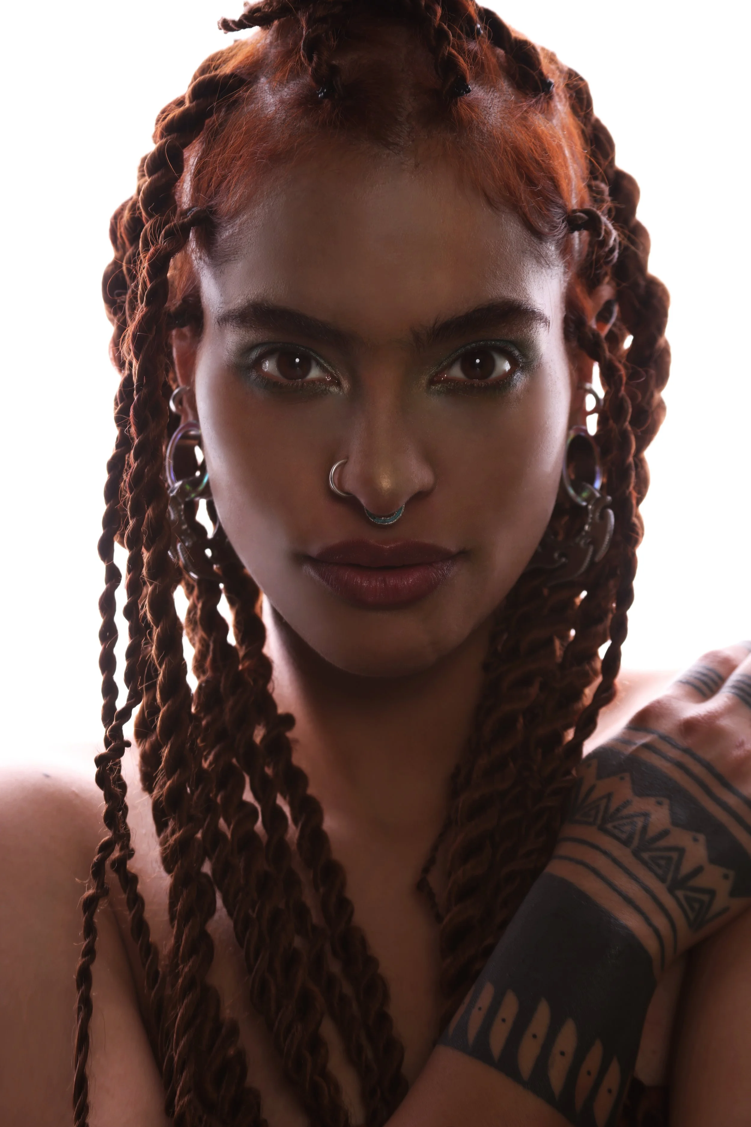 Close-up of a woman's face and upper body with brown dreadlocks, piercings on her nose and ears, and a tattoo on her arm, against a plain white background.
