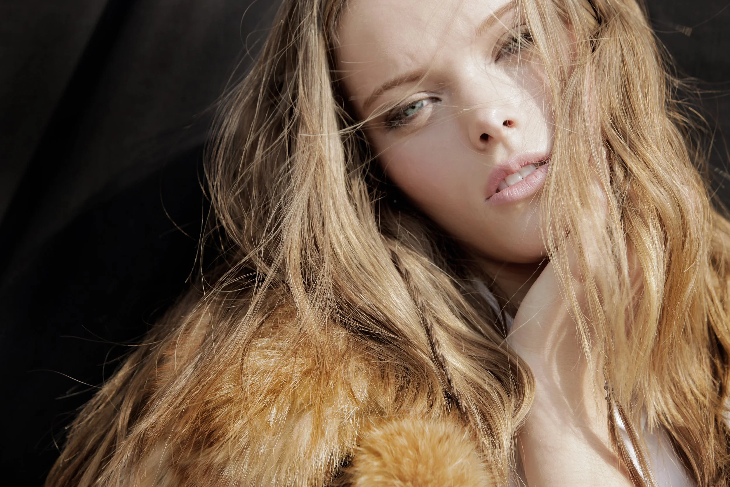 Close-up of a young woman with long, wavy, reddish-blonde hair and piercing blue eyes, with part of her face slightly tilted and one eye half-closed, against a dark background.