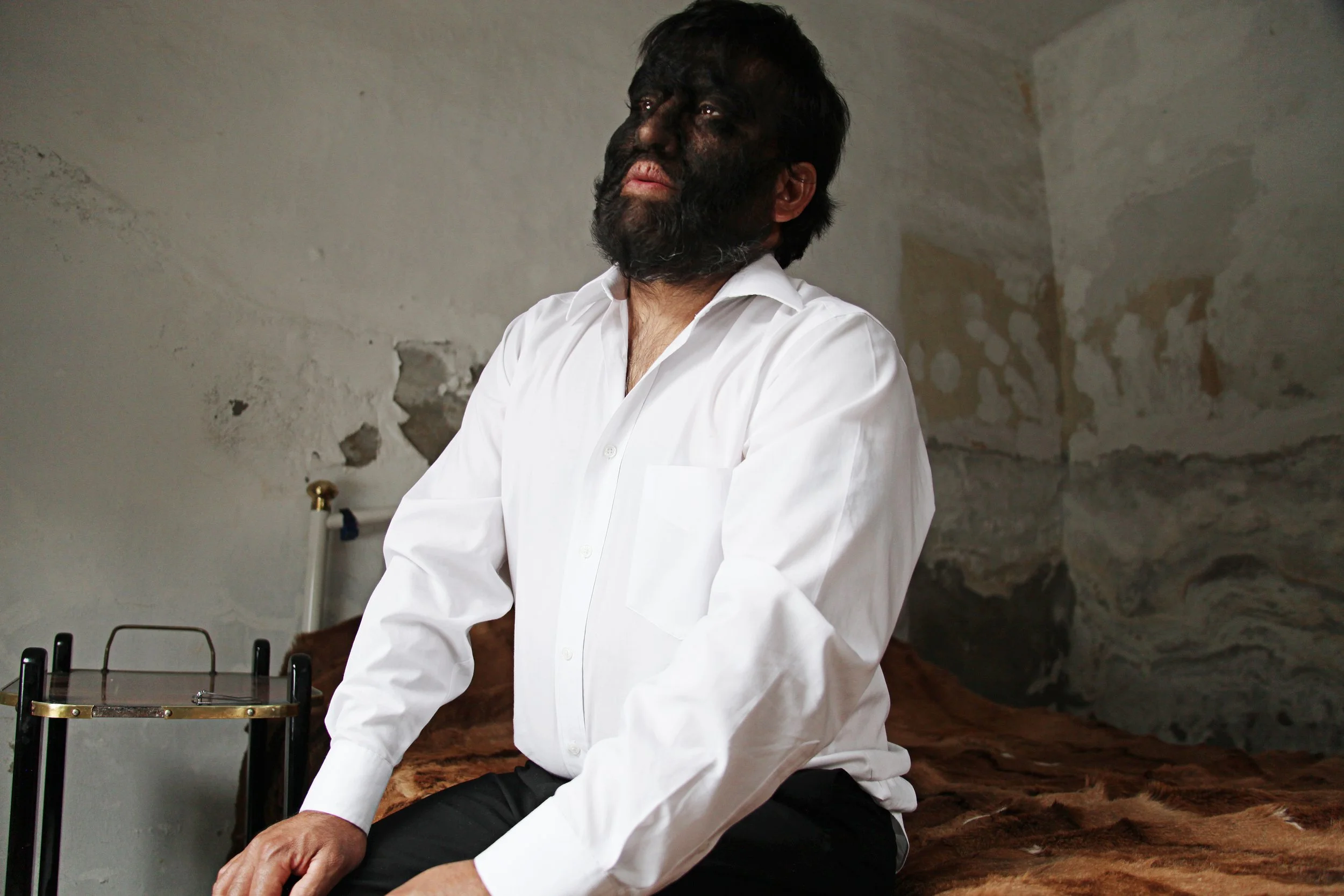A man with black hair and a beard wearing a white shirt, sitting cross-legged on a furry brown rug, with a plain, rough wall behind him and some furniture in the background.