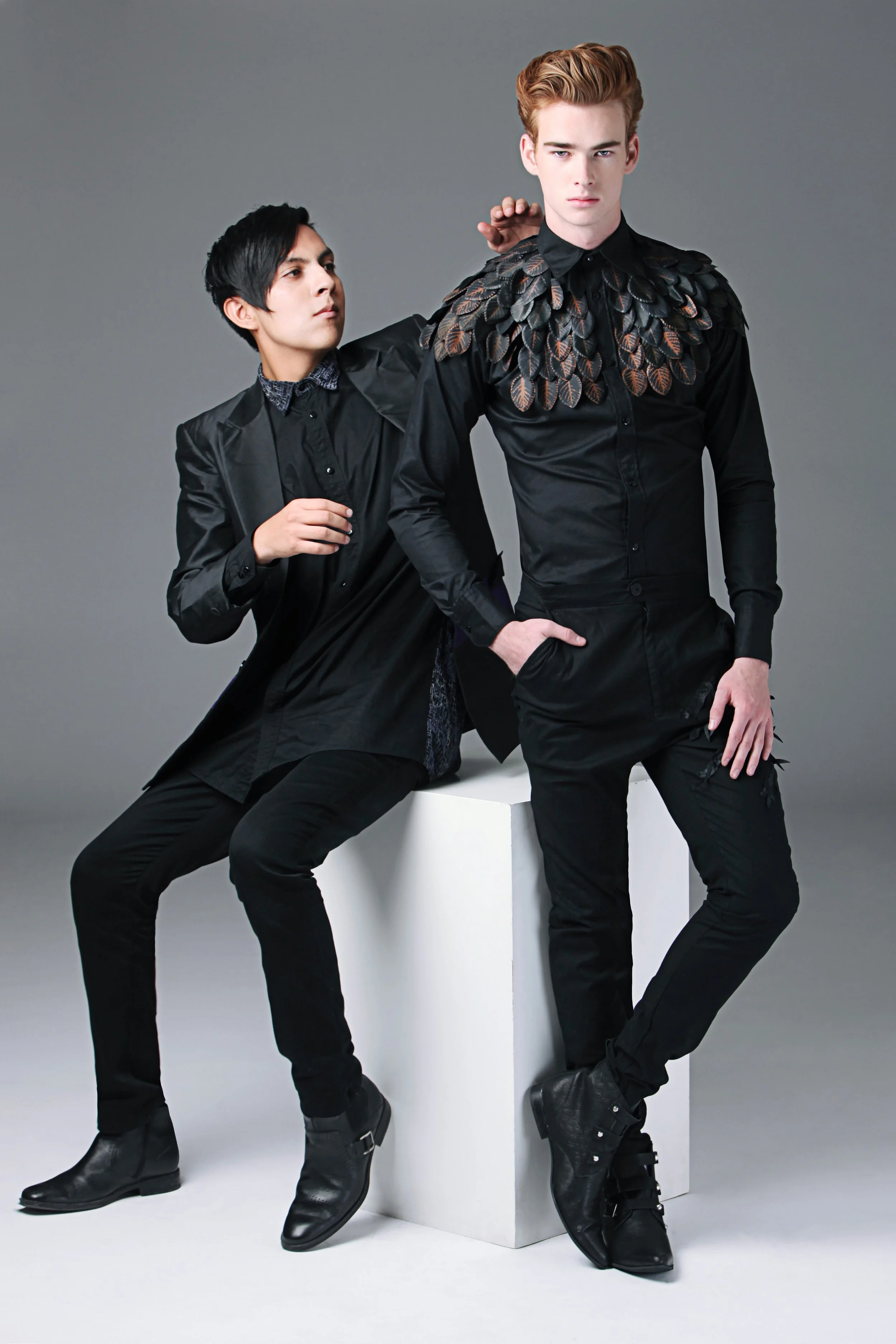 Two young men in fashionable black clothing and boots posing against a gray background, one sitting on a white block and the other standing with his hand on his hip.