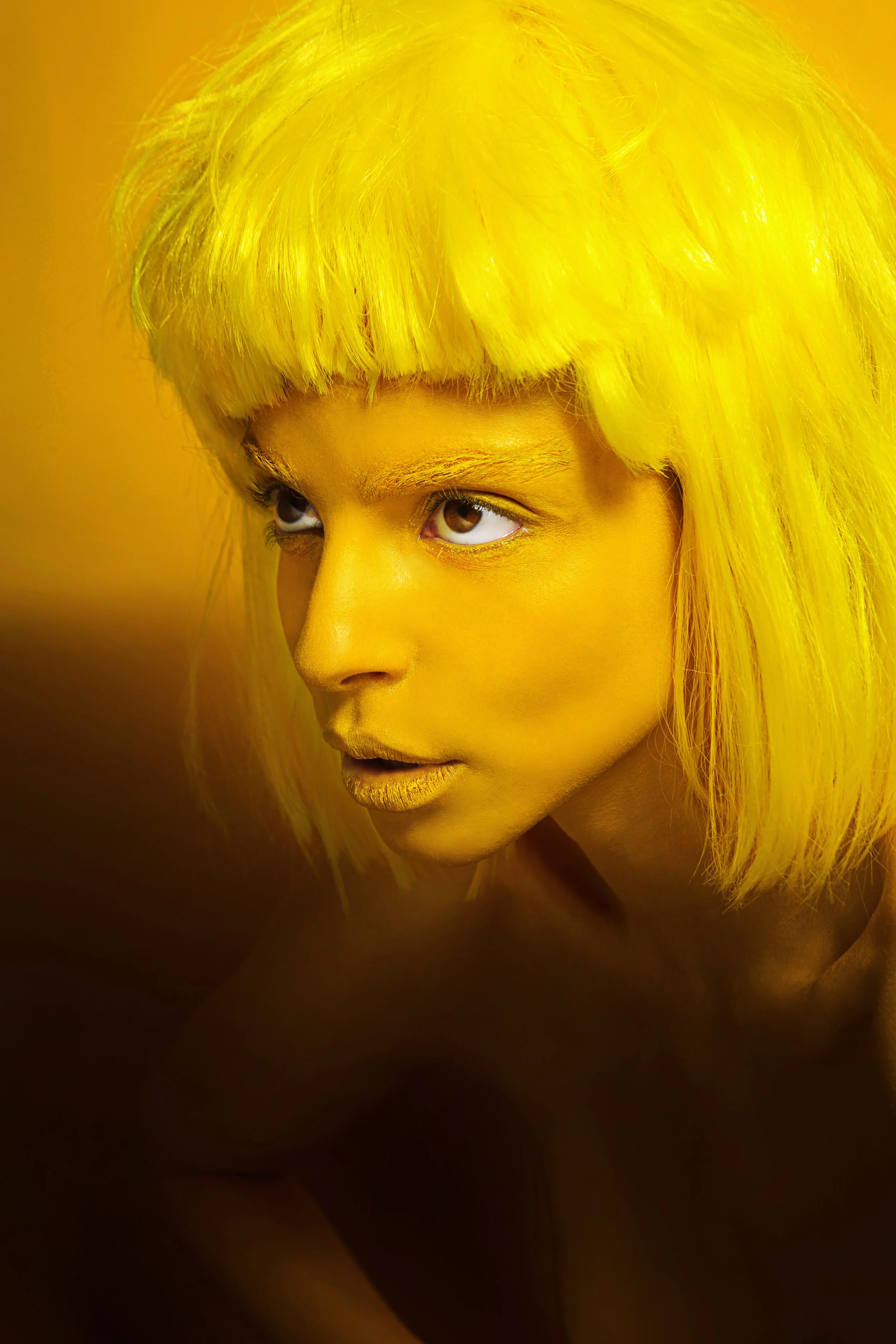 A close-up portrait of a person with vibrant yellow hair, yellow makeup, and a yellow background. The person is looking sideways, with brown eyes and golden lipstick.