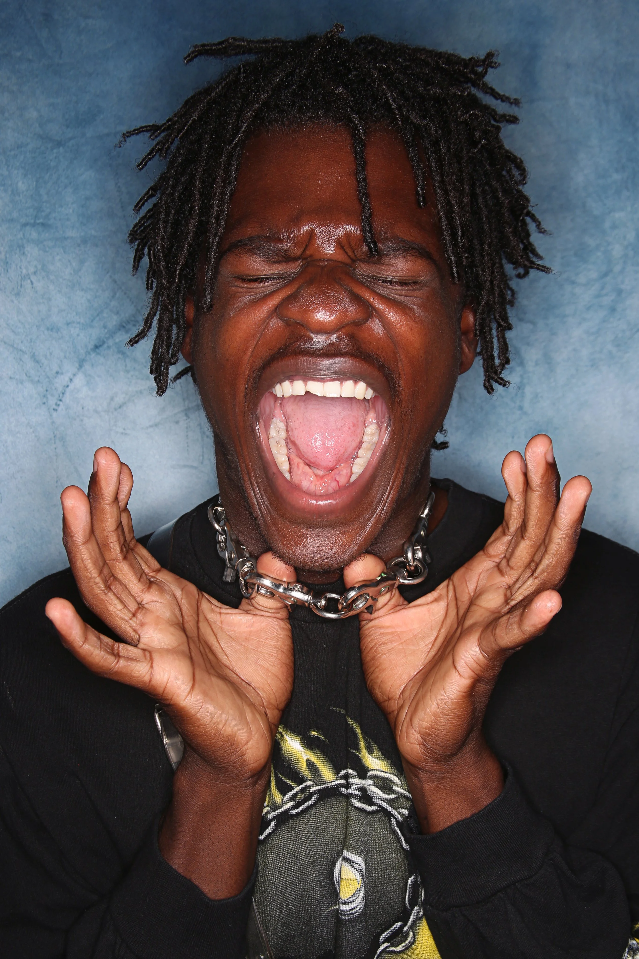 Person with dreadlocks and dark skin, screaming with mouth open wide, hands raised near face, against a blue background.