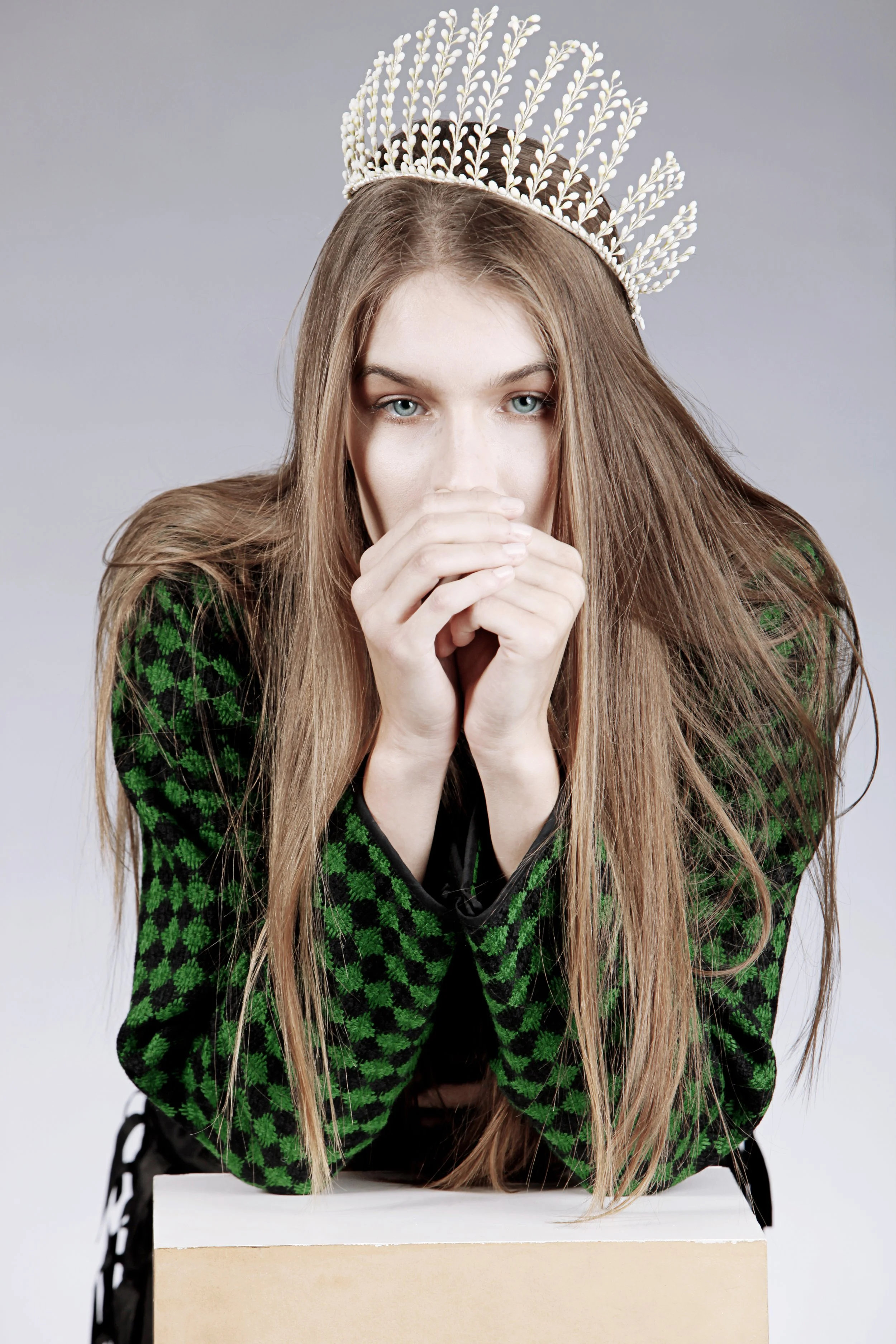 A woman with long, light brown hair wearing a white crown and a green and black patterned sweater, sitting behind a beige table, with her hands covering her mouth and chin, against a plain gray background.