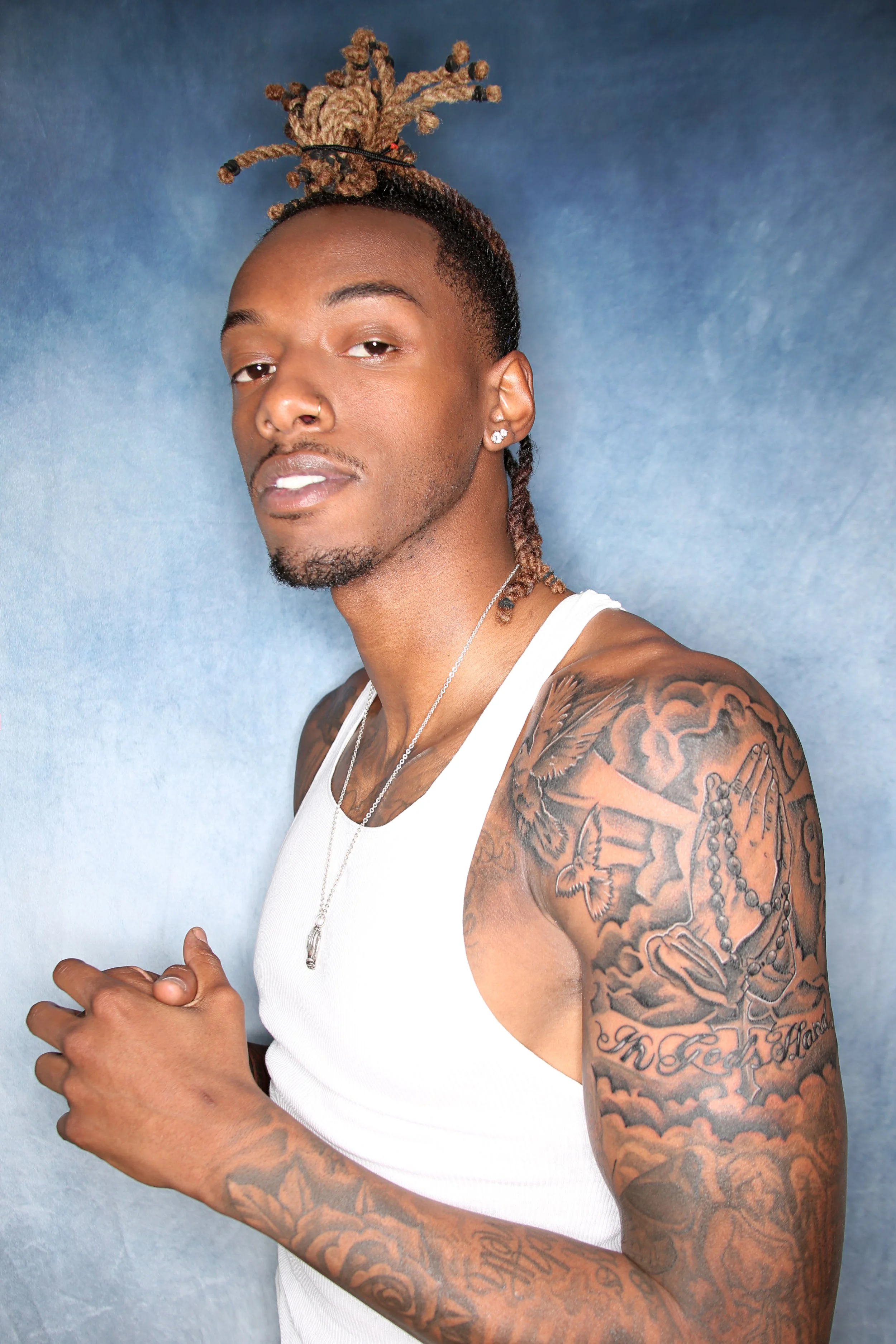 A man with a fashionably styled updo hairstyle, tattoos on his arm, wearing a white tank top, earrings, and a silver necklace, posing against a blue textured background.