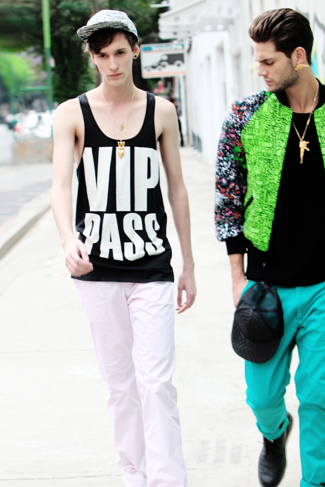 Two young men walking outdoors in an urban area. One is wearing a sleeveless black shirt with big white text saying 'VIP PASS', pink pants, and a patterned cap. The other is wearing a colorful jacket, turquoise pants, and black sneakers.