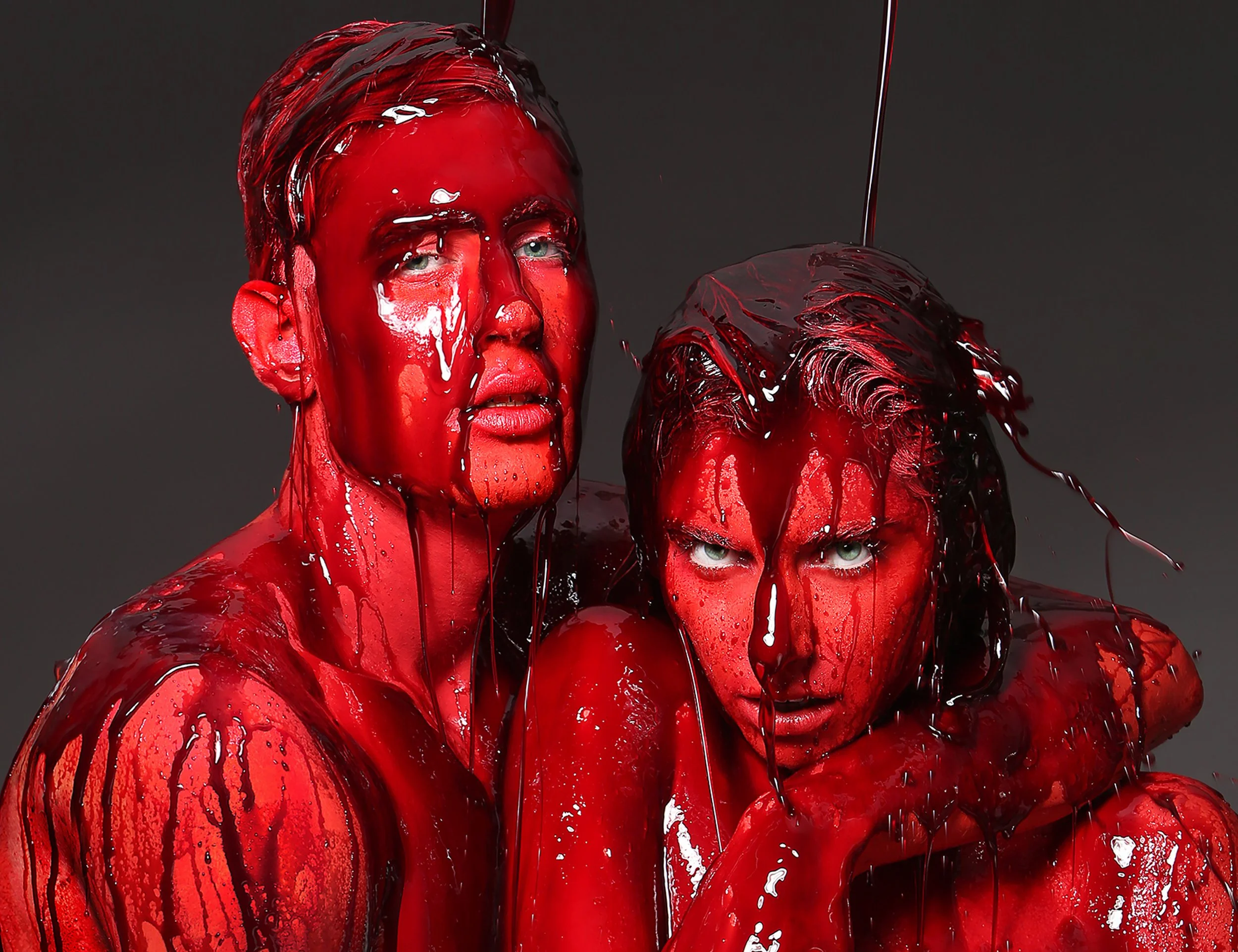 Portrait of three people covered in red paint, with paint dripping down their faces and bodies, against a dark background.