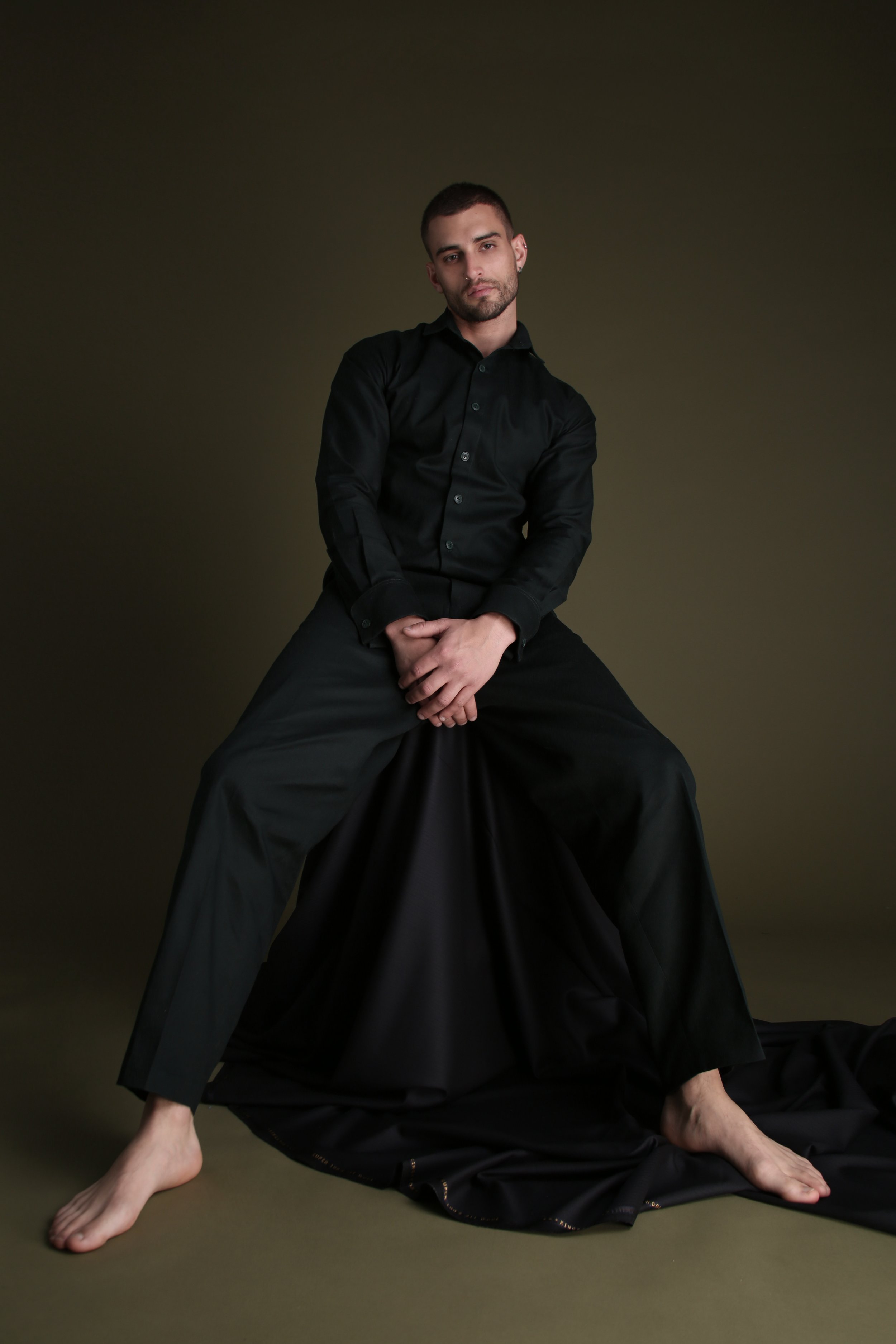 A young man with short dark hair and a beard, sitting on the floor with only his right foot touching the ground, wearing a black long-sleeve shirt and black pants against a dark green background.