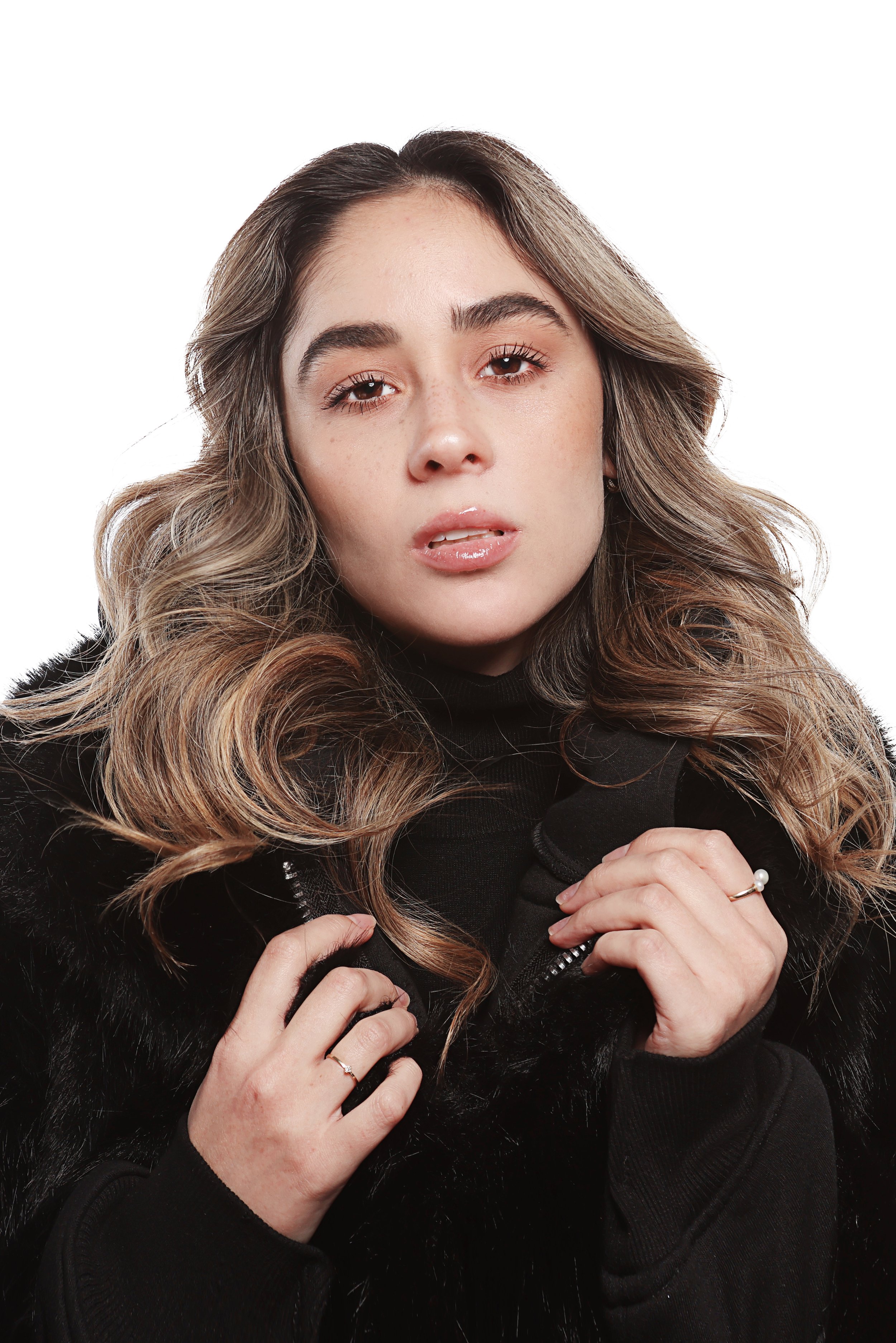 A woman with long wavy hair, wearing a black jacket, poses with a serious expression, holding the zipper of her jacket with both hands.