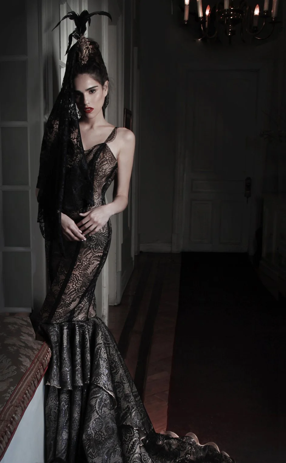 A woman dressed in an elegant, black lace gown with a mermaid silhouette, standing in a dimly lit room with classical decor and a chandelier. She has an elaborate headpiece with feathers and lace veil.