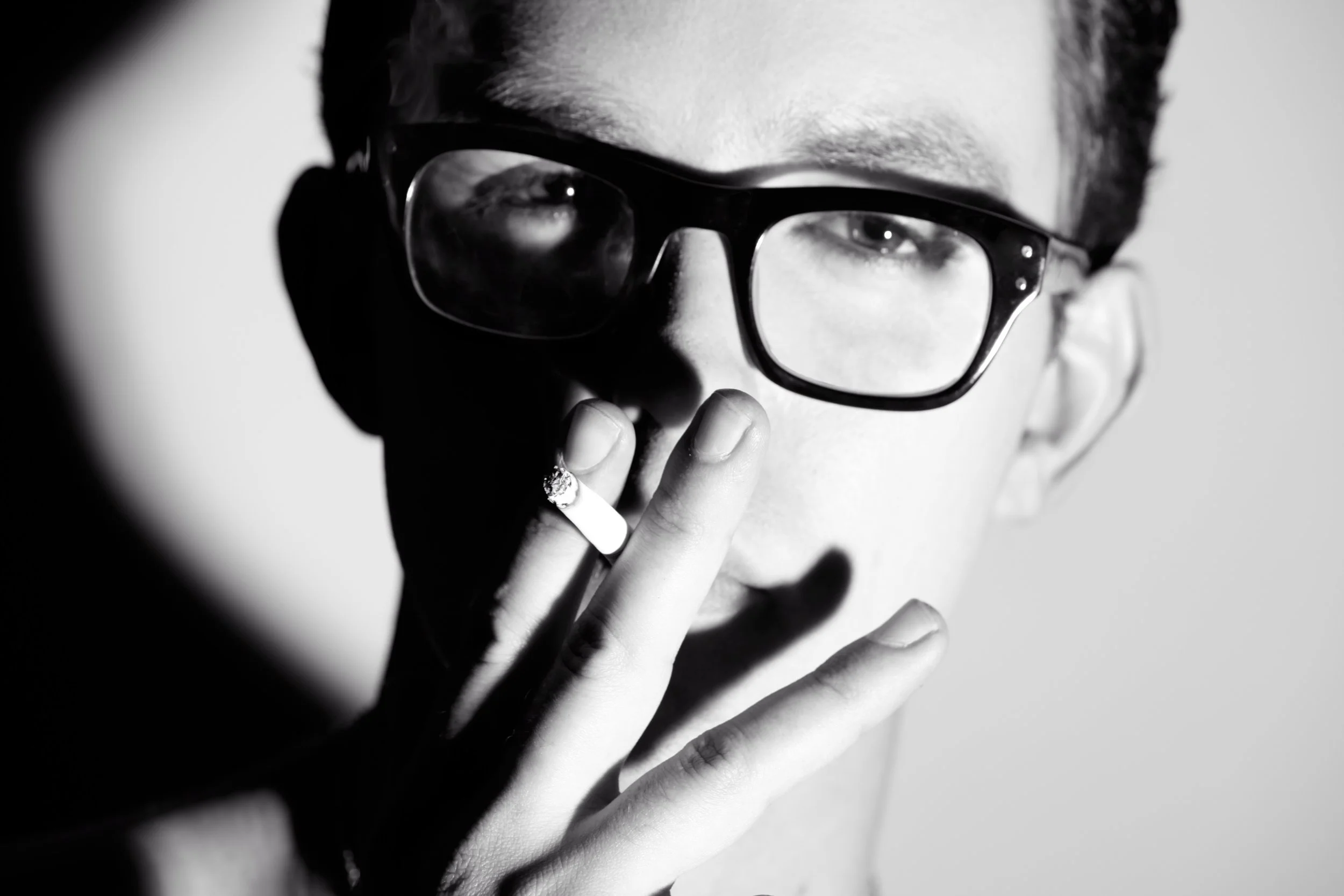 A black and white close-up photograph of a person with a hand near their face, wearing glasses, and smoking a cigarette.