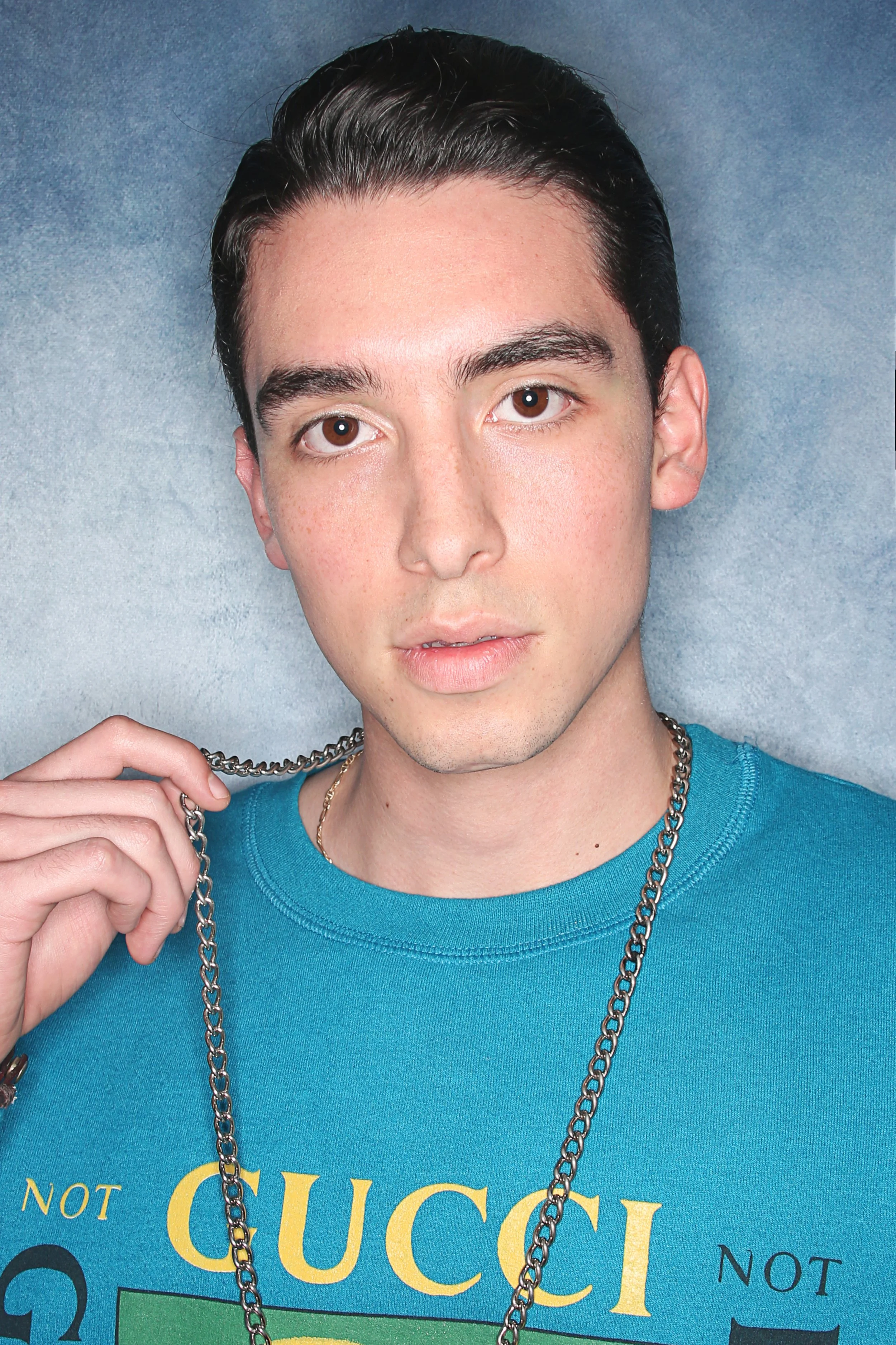 Close-up of a young man with dark, slicked-back hair, light skin with freckles, wearing a blue T-shirt and multiple chains, holding a chain in his hand, against a plain, smoke-colored background.