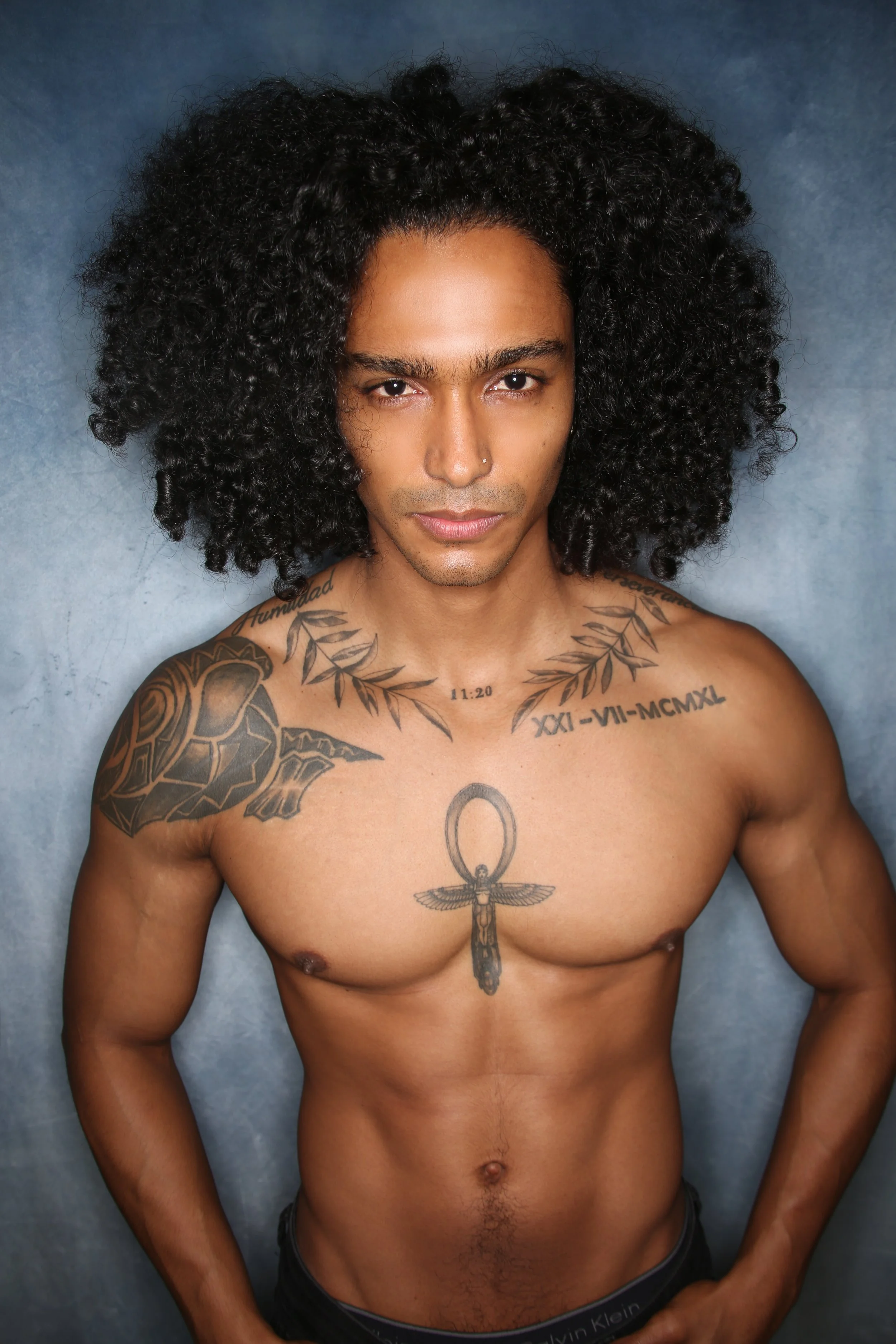 A shirtless man with dark, curly hair and tattoos on his chest and arms, standing against a gray background.