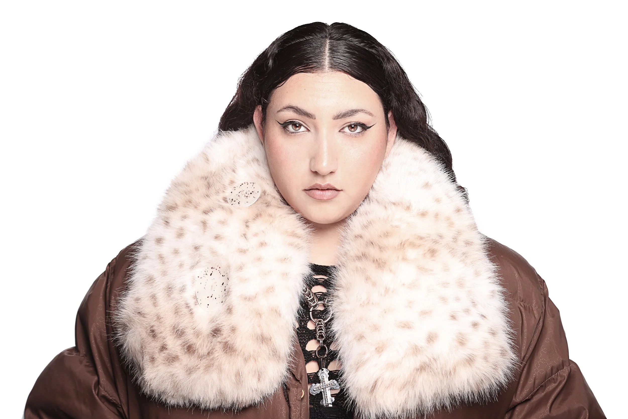 A woman with dark hair and eyeliner, wearing a brown jacket with a large faux fur collar, standing against a plain white background.