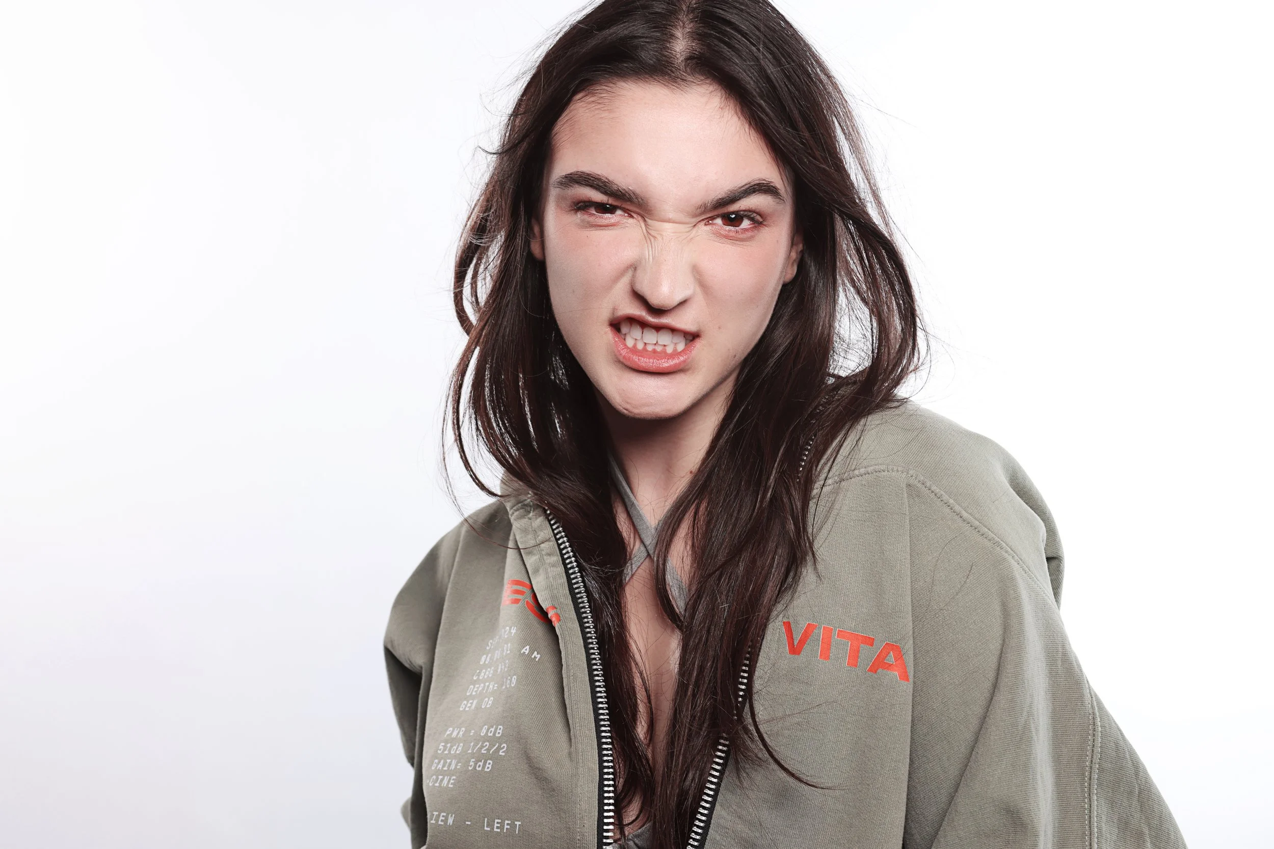 A young woman with long dark hair making an angry or fierce facial expression, scrunching her eyebrows, showing her teeth, and looking directly at the camera. She is wearing a green zip-up jacket with red and white lettering on the chest, against a p