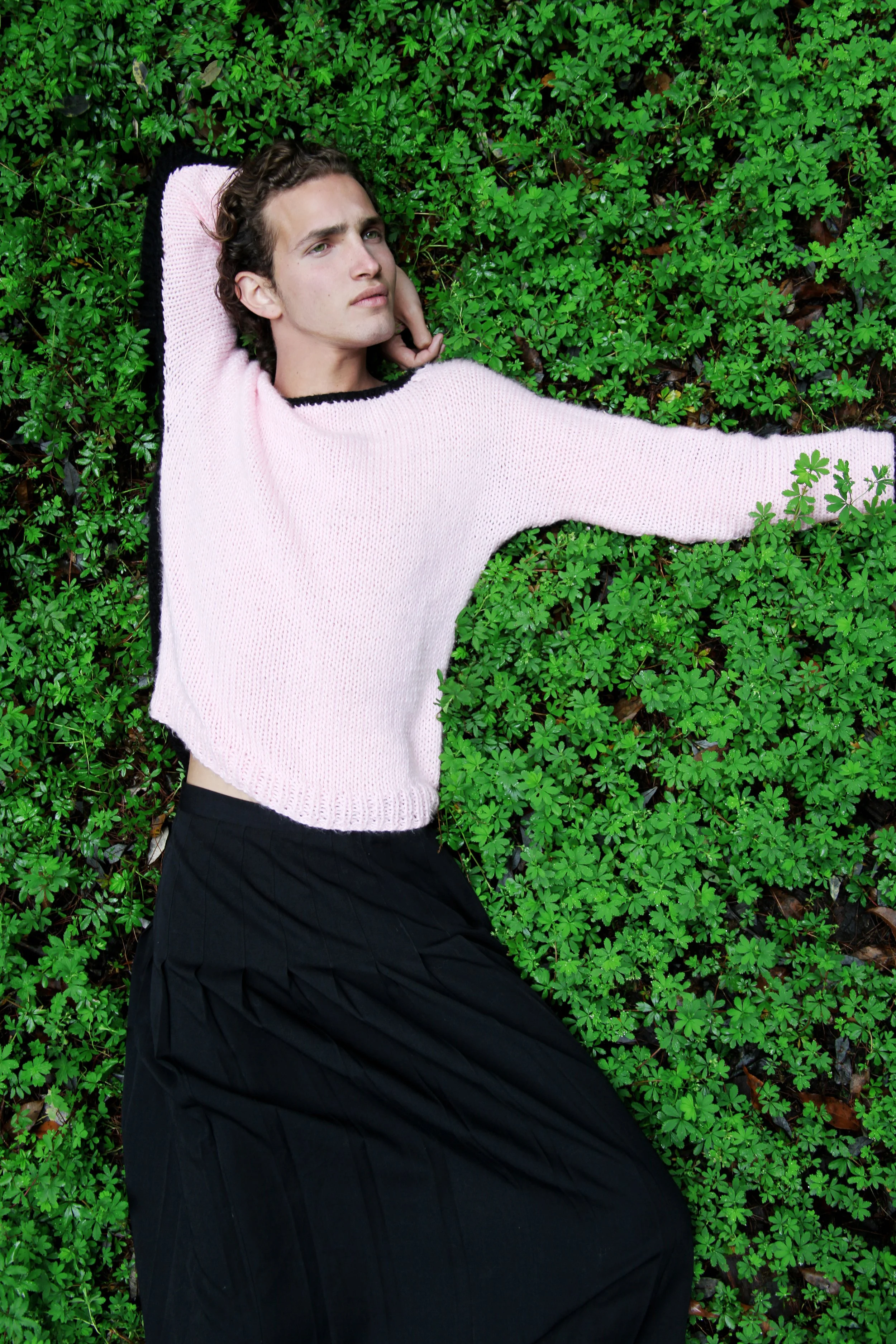 A young man lying on a bed of green clover leaves, wearing a pink sweater and black skirt, gazing contemplatively into the distance.