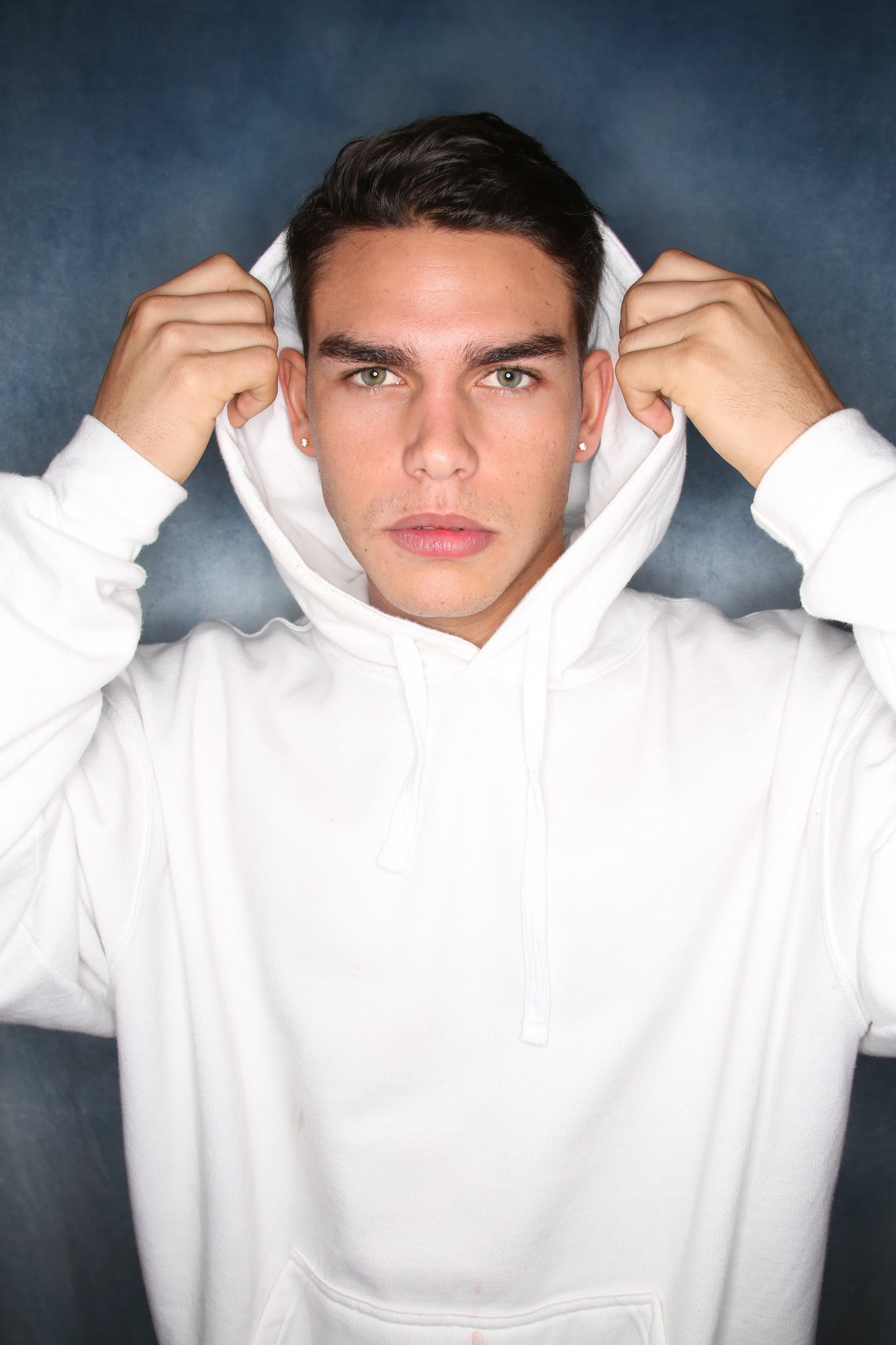 A young man with short dark hair and light skin is pulling up the hood of his white hoodie and looking directly at the camera. He has green eyes, small earrings, and a neutral expression. The background is a dark, textured surface.