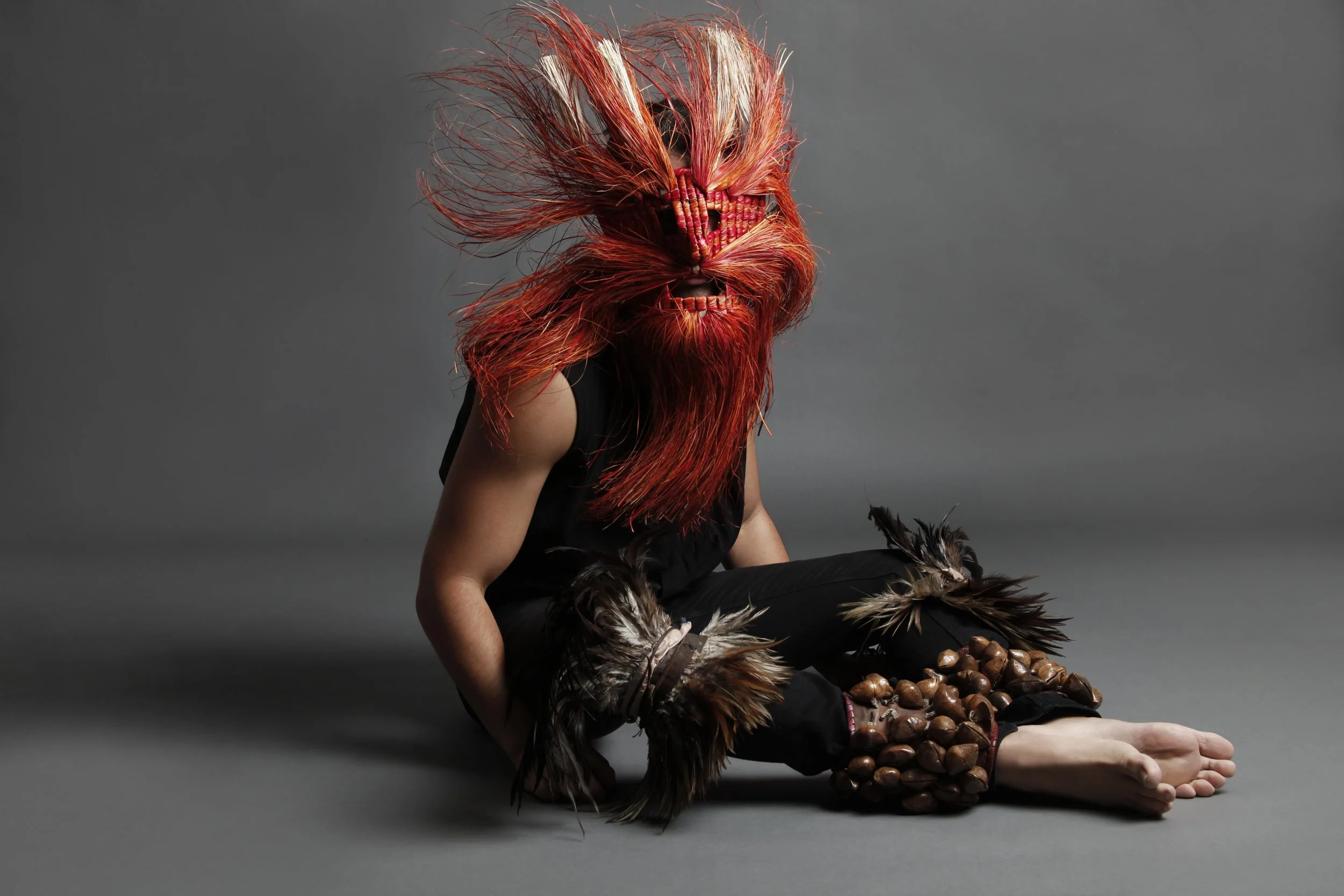 Person wearing an elaborate animal-inspired mask and costume, sitting on the floor with legs crossed, against a plain gray background.