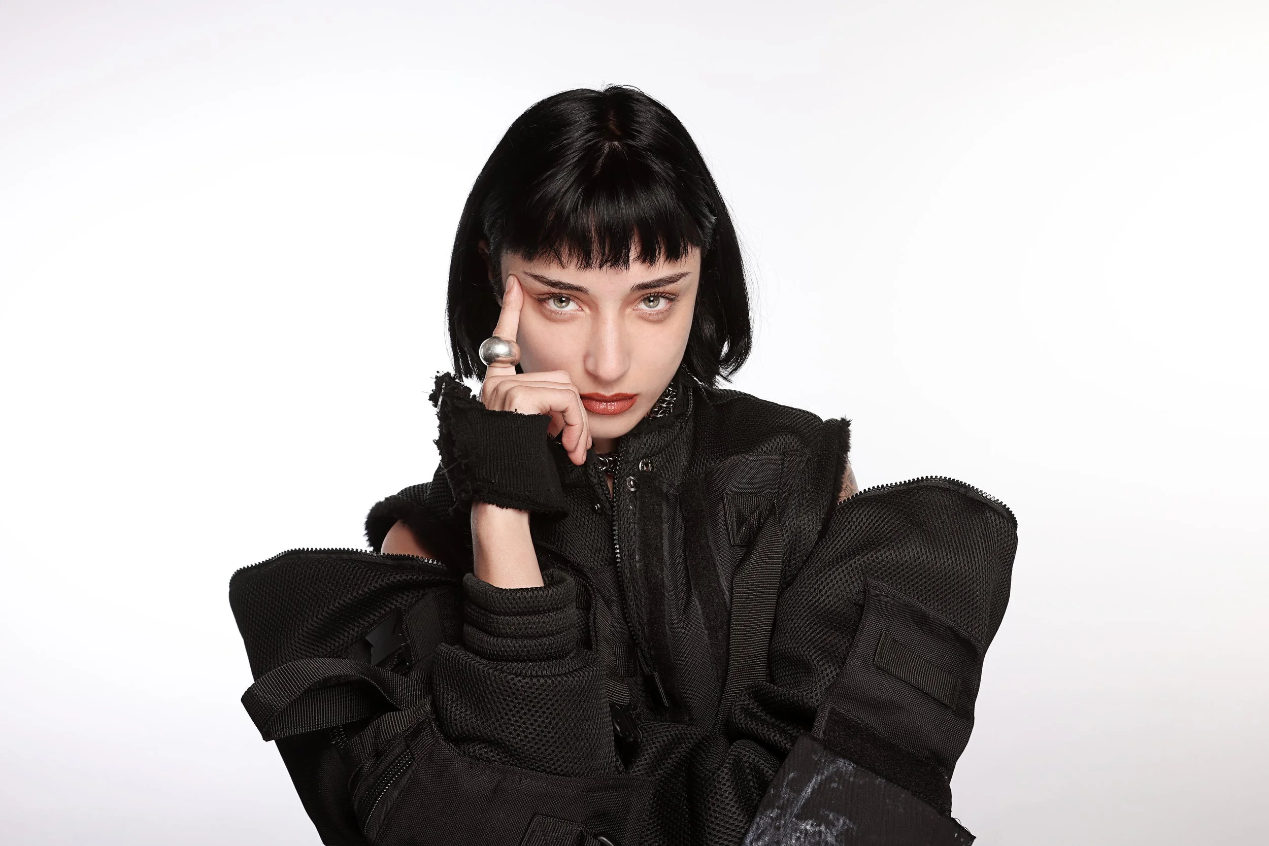 A woman with short black hair and bangs, wearing a black jacket with zippers, is posing with her right hand touching her forehead and her left arm crossed over her lap, against a plain white background.