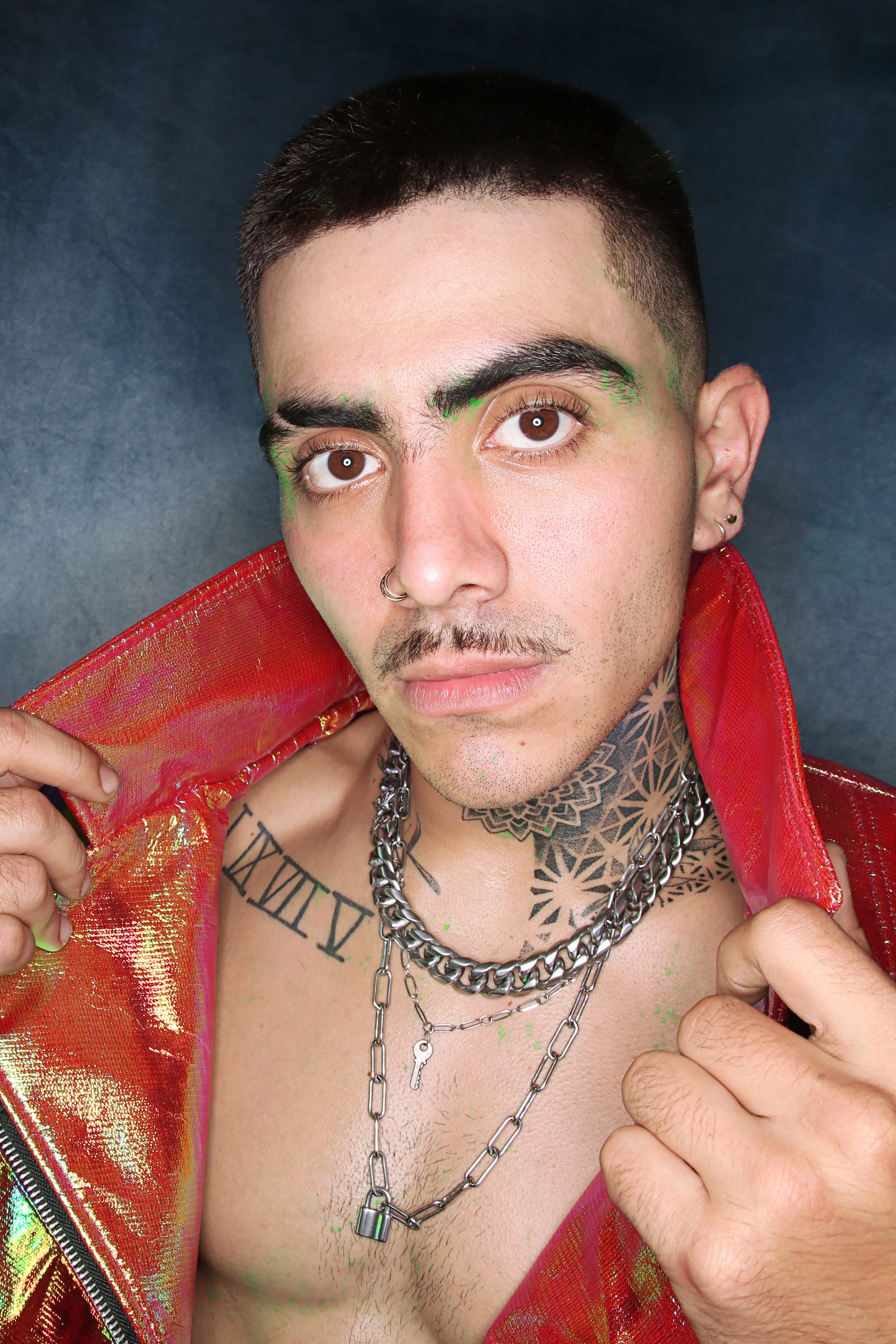 Close-up of a young man with a short haircut, brown eyes, and multiple facial piercings. He is wearing layered silver chains and has tattoos on his neck and chest. He is wearing a shiny red and gold jacket, slightly open to reveal chest hair. His exp