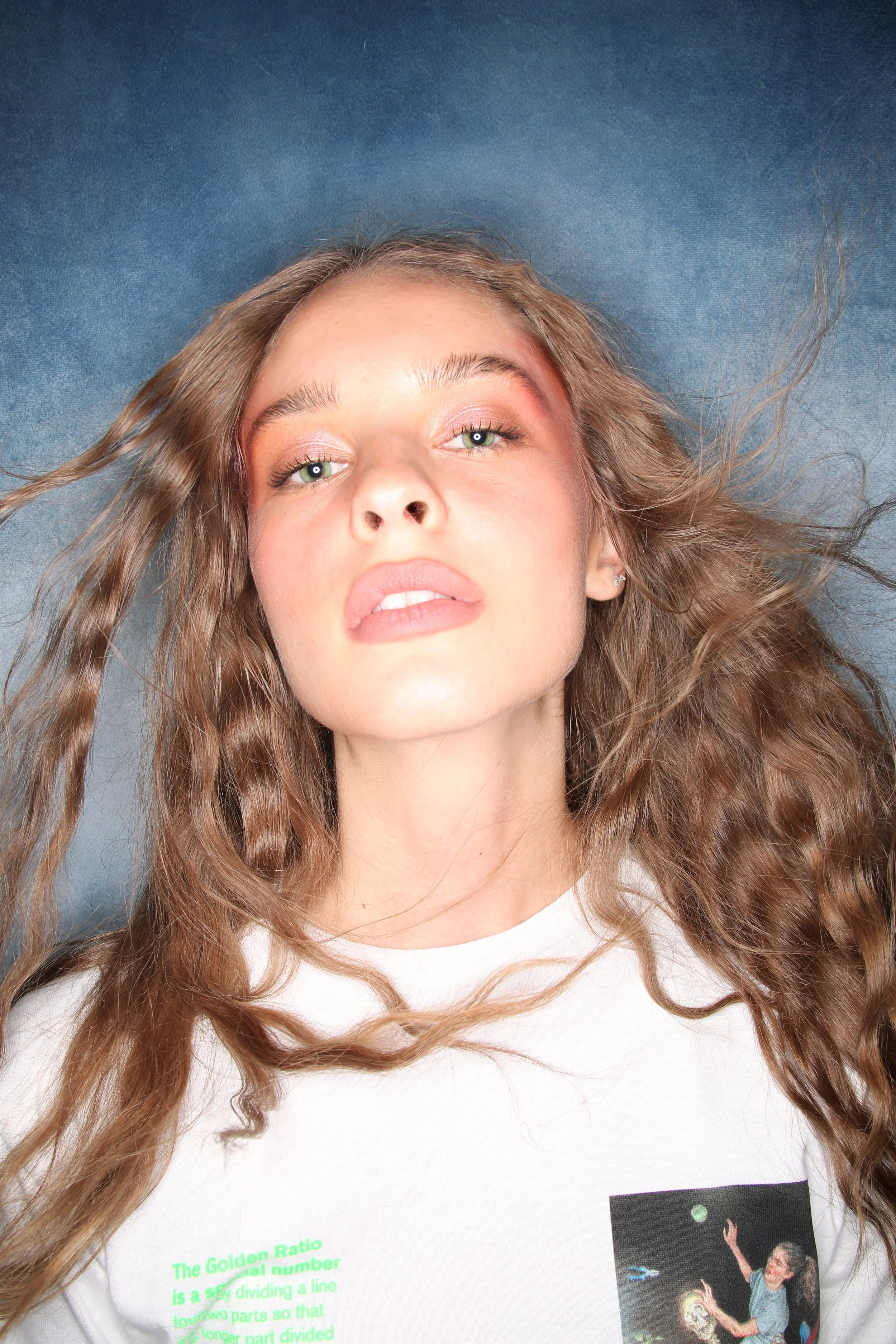 A woman with long curly hair lying down on a blue background, wearing a white shirt with a graphic and green text.