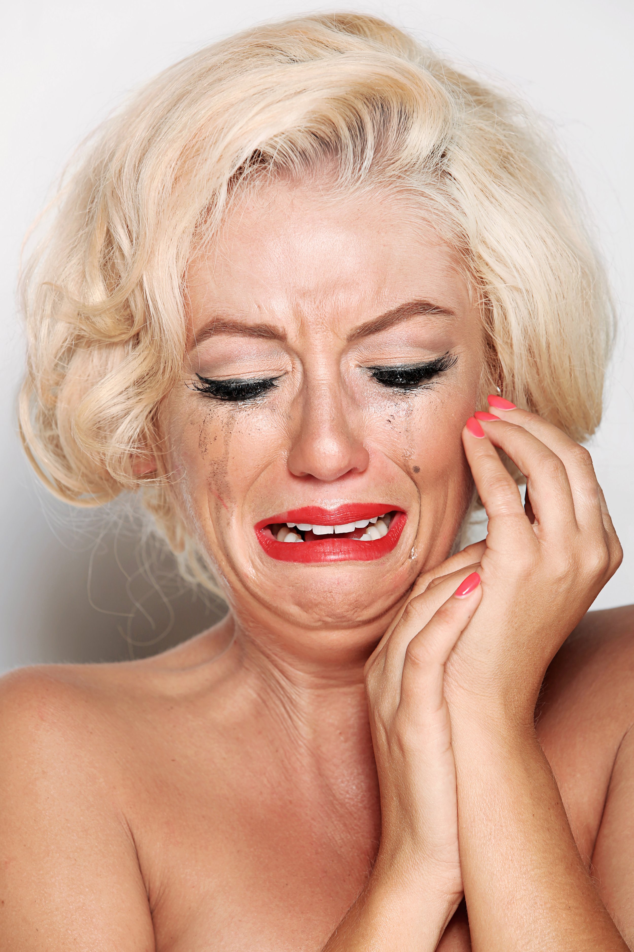 A close-up of a woman with platinum blonde hair and light skin showing tears and an emotional expression. She has red lipstick, black eyeliner, and manicured nails, with her hand touching her face.