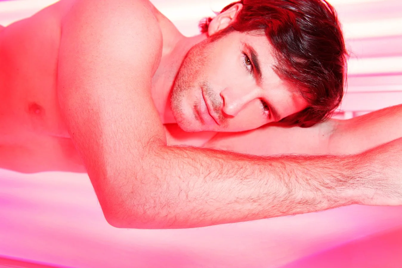 A shirtless man with brown hair and stubble lying on his side on a pink surface, looking at the camera with a relaxed expression.
