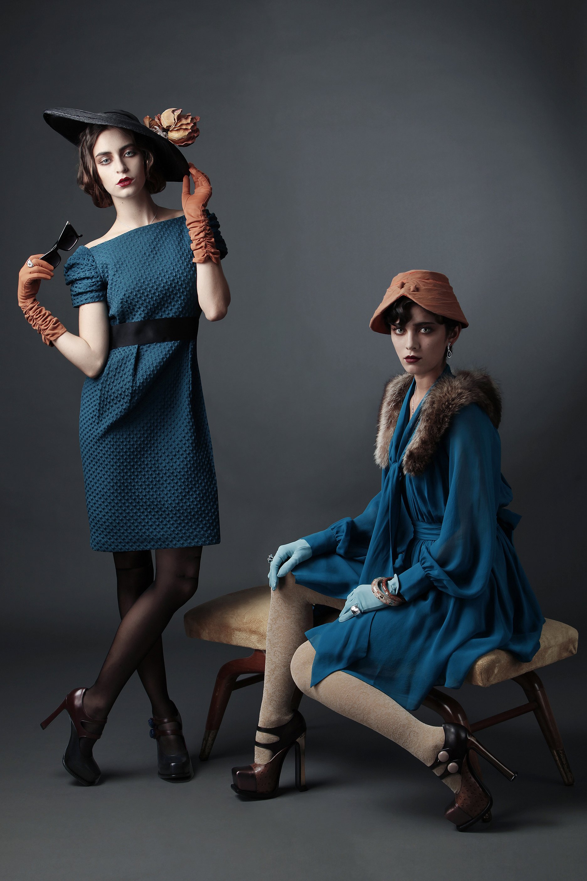 Two women in vintage-style fashion posing against a gray background. One standing in a blue dress with puffed sleeves, wearing a large black hat with a flower, holding sunglasses, and wearing orange gloves. The other sitting on a wooden chair in a bl