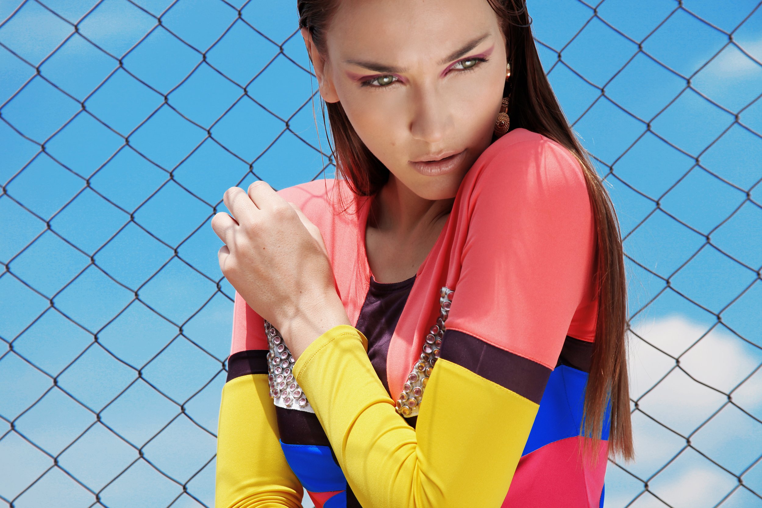 A woman with green eyes and long brown hair wearing a colorful outfit with pink, yellow, and blue sections, accessorized with large earrings and beaded necklaces, standing in front of a chain-link fence against a blue sky.