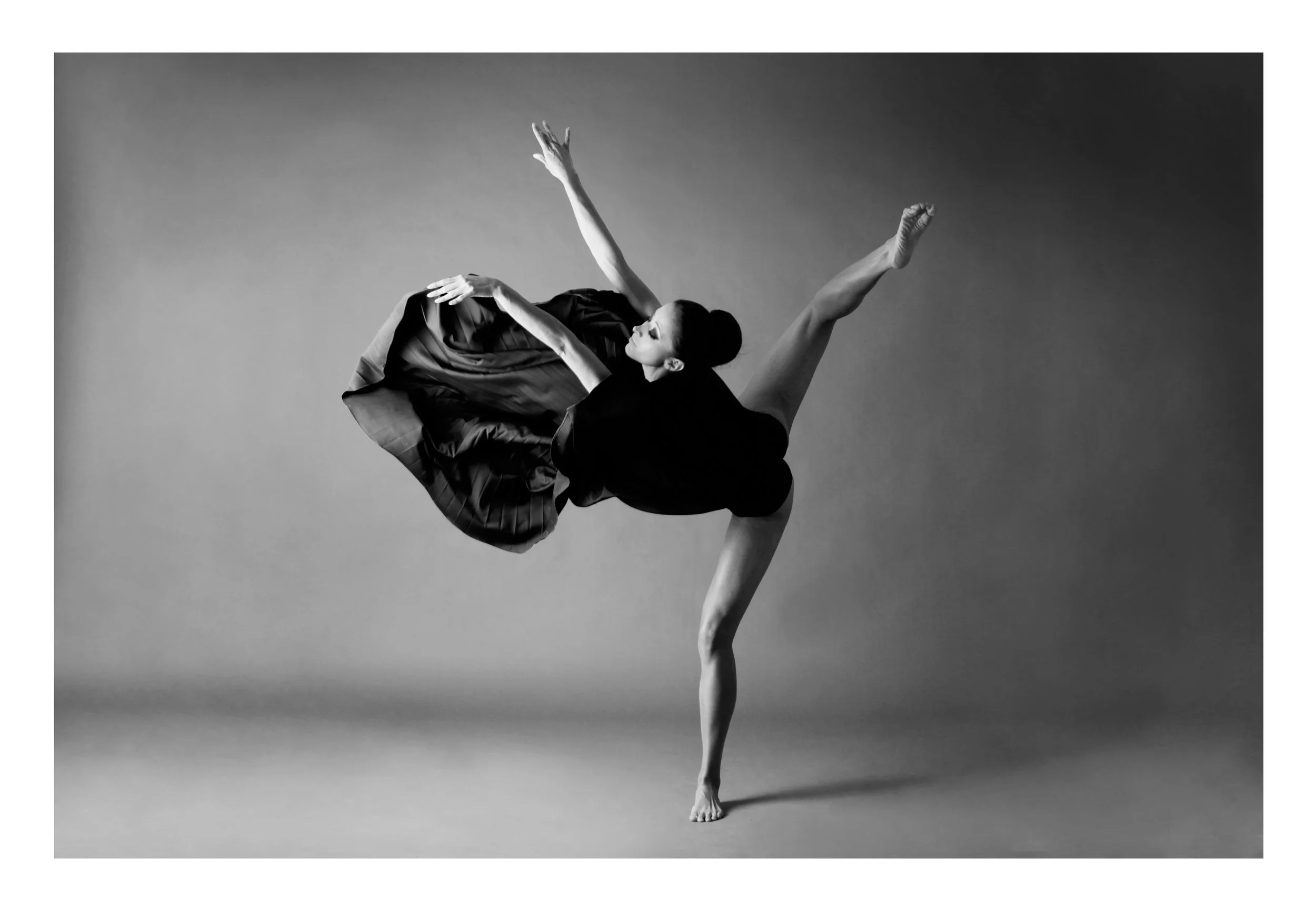 A dancer in a black dress balancing on one foot with arms raised and one leg extended backward, captured in a black and white photograph.