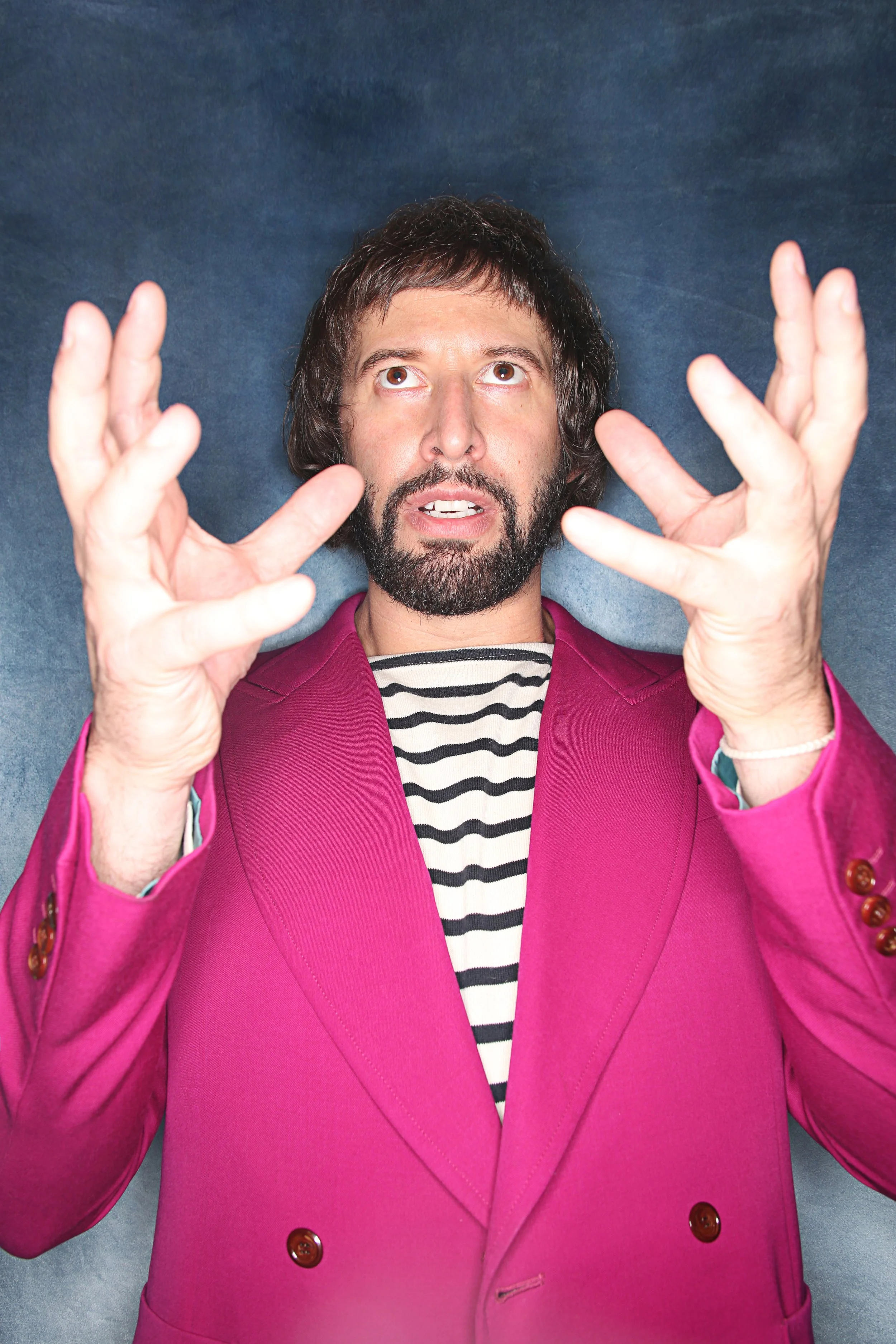 Man with a beard and dark brown hair looking up, wearing a bright pink blazer, striped shirt, and light-colored background.