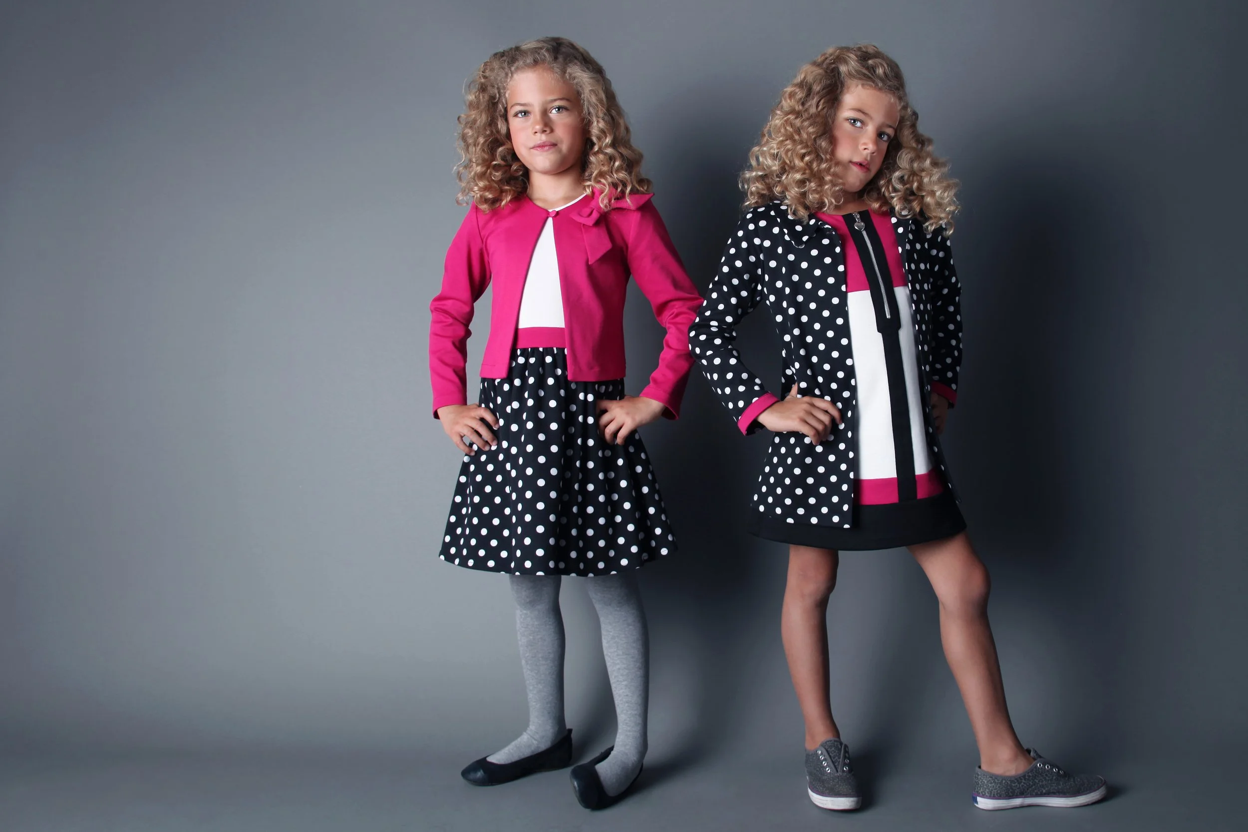 Two young girls with curly hair wearing polka dot skirts and colorful jackets, standing against a plain gray background.