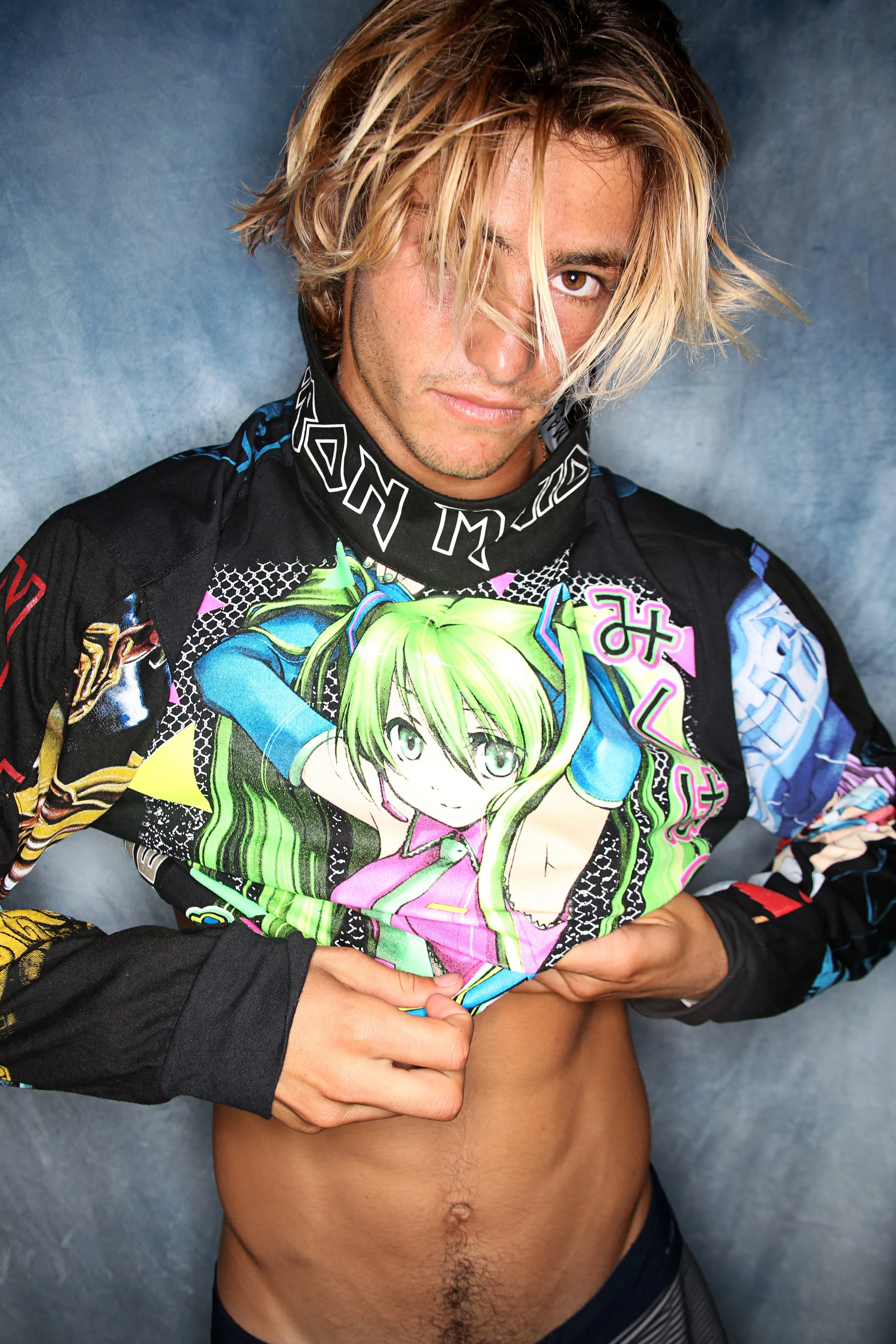 A man with shoulder-length blond hair, brown eyes, and a light complexion is holding a colorful anime print with a green-haired girl. He is wearing a racing suit with Japanese characters and graphic designs, and is posing against a plain gray backgro