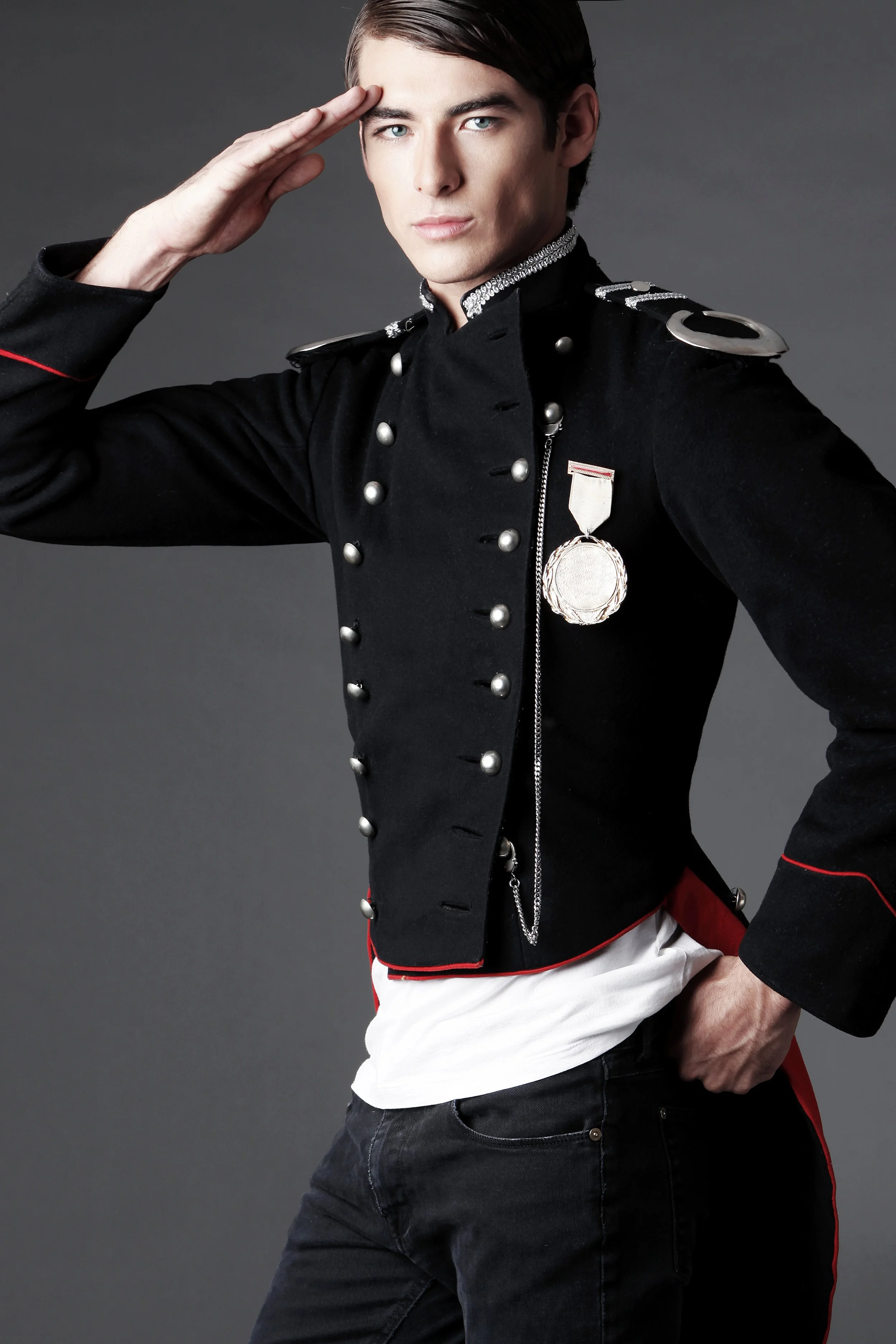 A young man in a military-style black jacket with silver buttons and medals, saluting with his right hand, standing against a plain gray background.