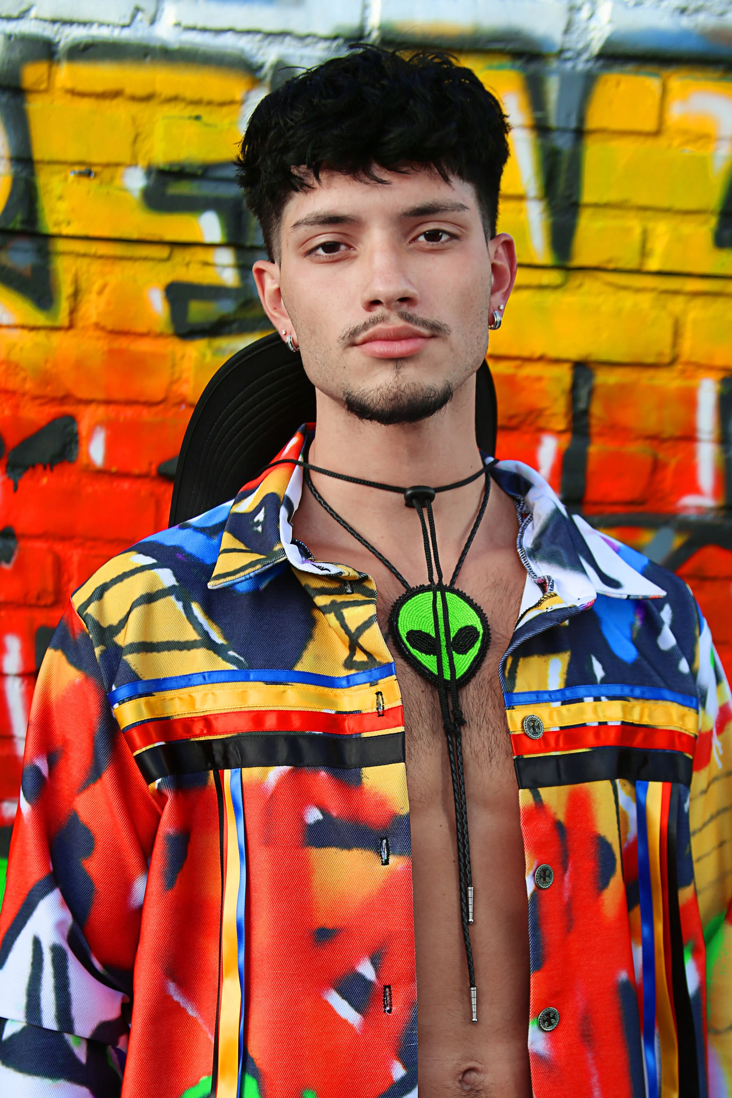 A young man with black curly hair, earrings, and a mustache, wearing a colorful, patterned shirt unbuttoned at the middle, stands in front of a graffiti-covered brick wall.