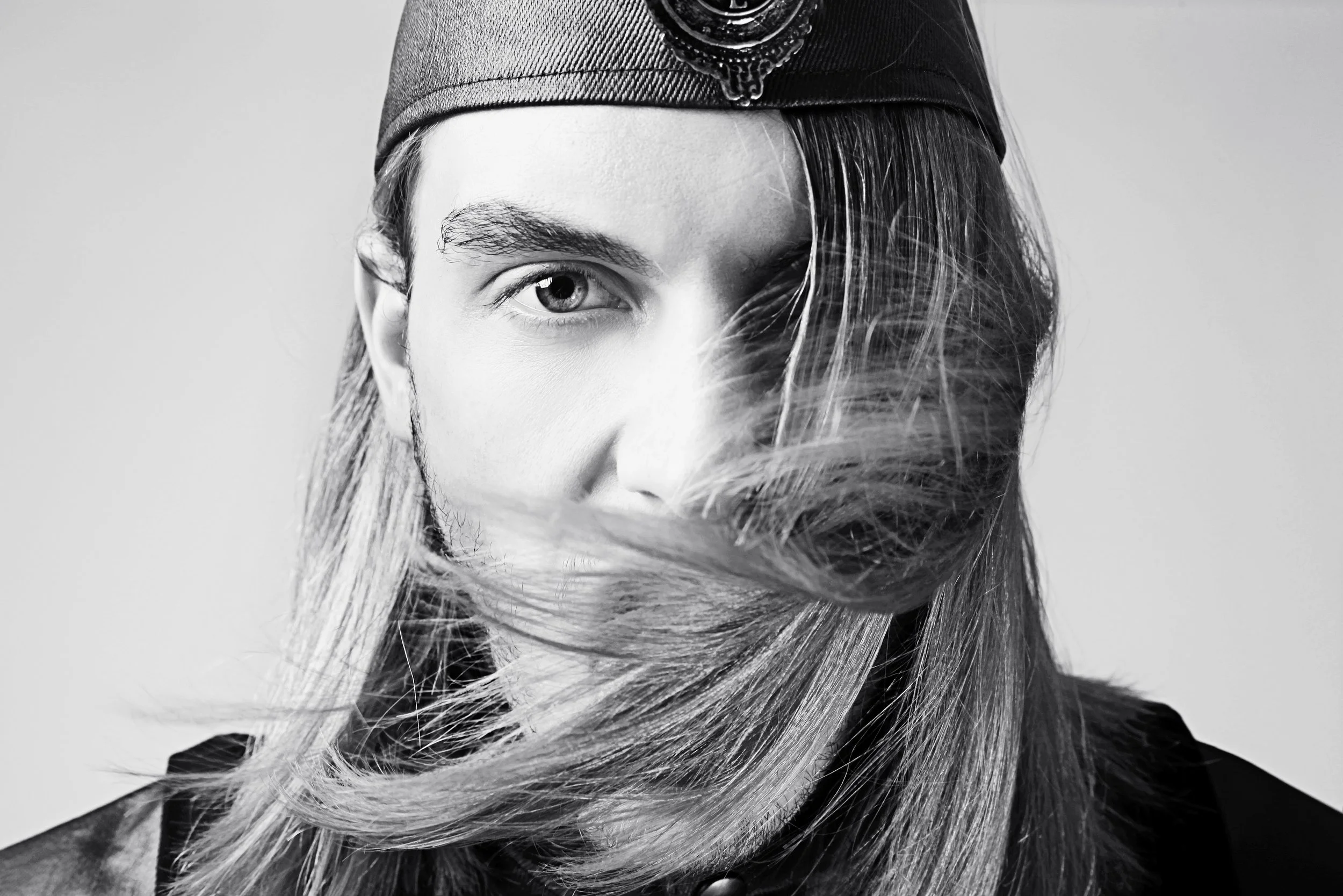 A black and white close-up portrait of a young woman wearing a cap, with long hair flowing around her face and partially obscuring one eye.