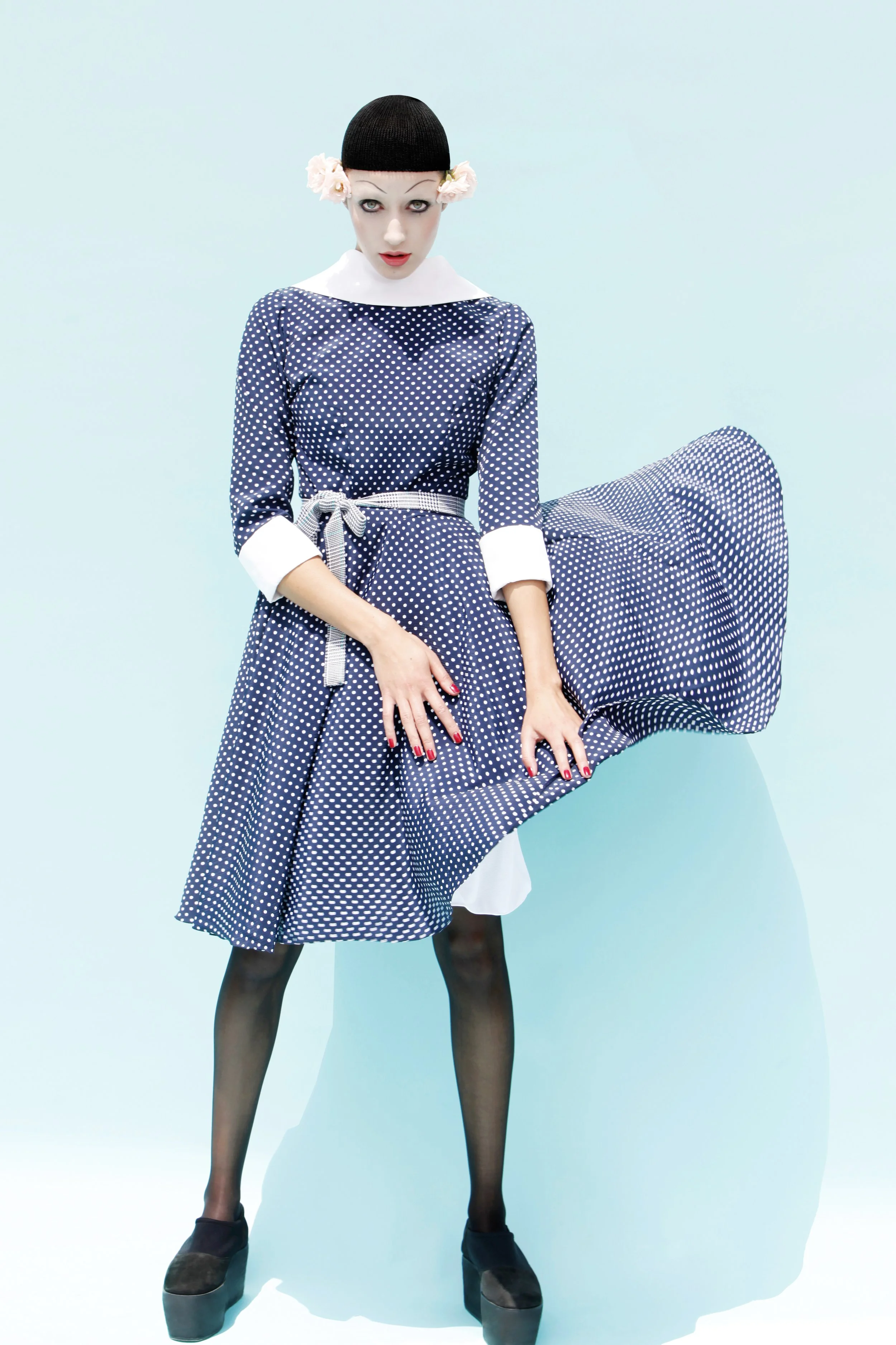 A woman with white face makeup, black wig cap with pink flowers, wearing a blue and white polka dot dress with white collar and cuffs, sheer black stockings, black platform shoes, against a light blue background.