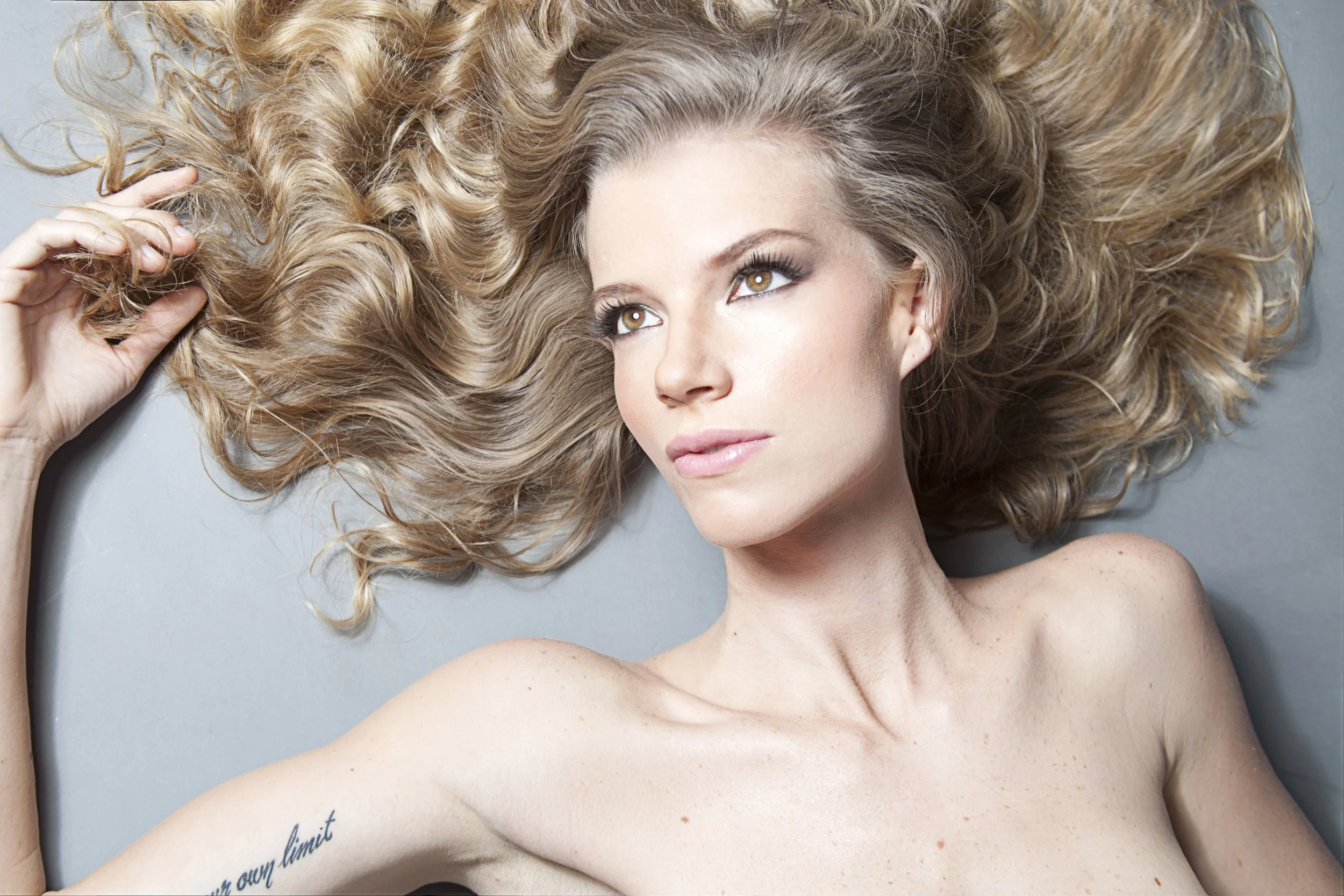 A woman with curly blonde hair lying on a gray surface, looking upward with a relaxed expression. She has makeup with defined eyes and pink lips, and a tattoo on her right side near her ribcage.
