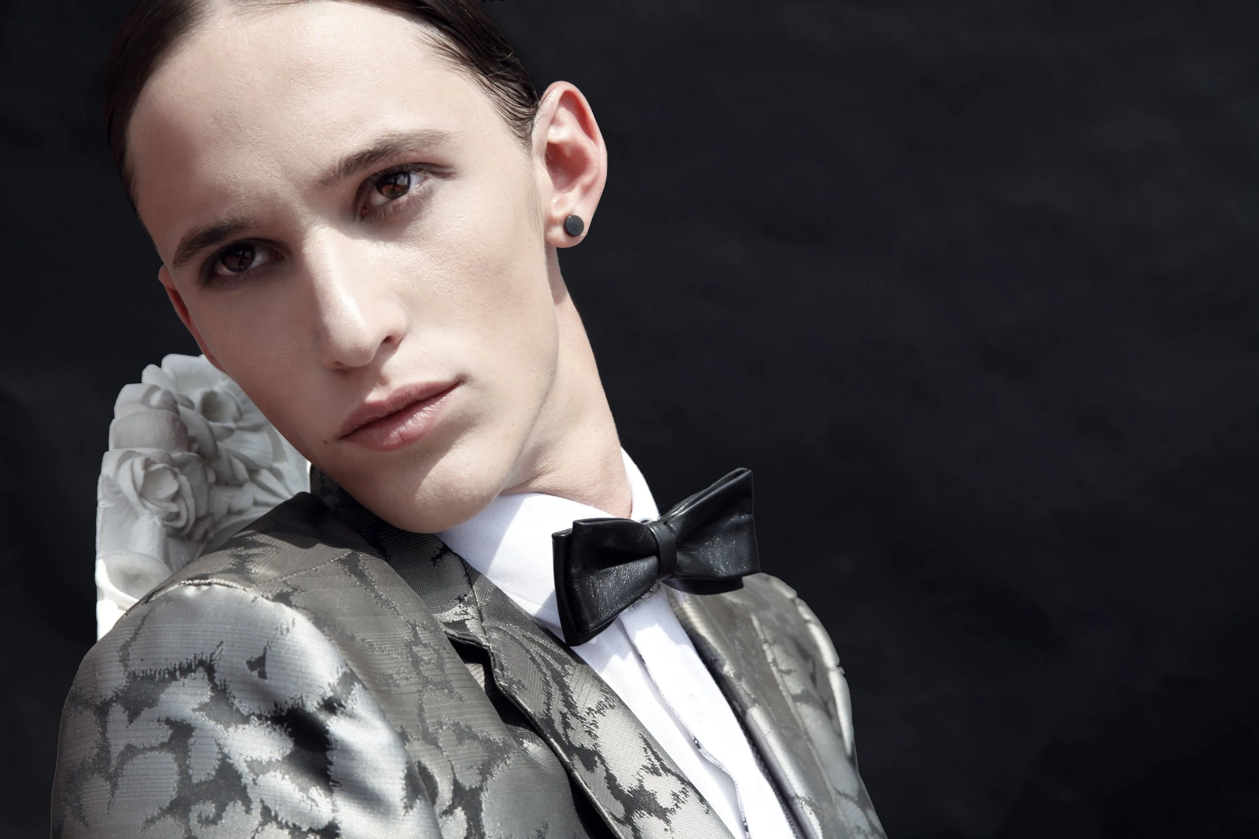 A woman with short dark hair and makeup wearing a patterned silver suit, white shirt, black bow tie, and a white flower accessory on her shoulder, against a black background.