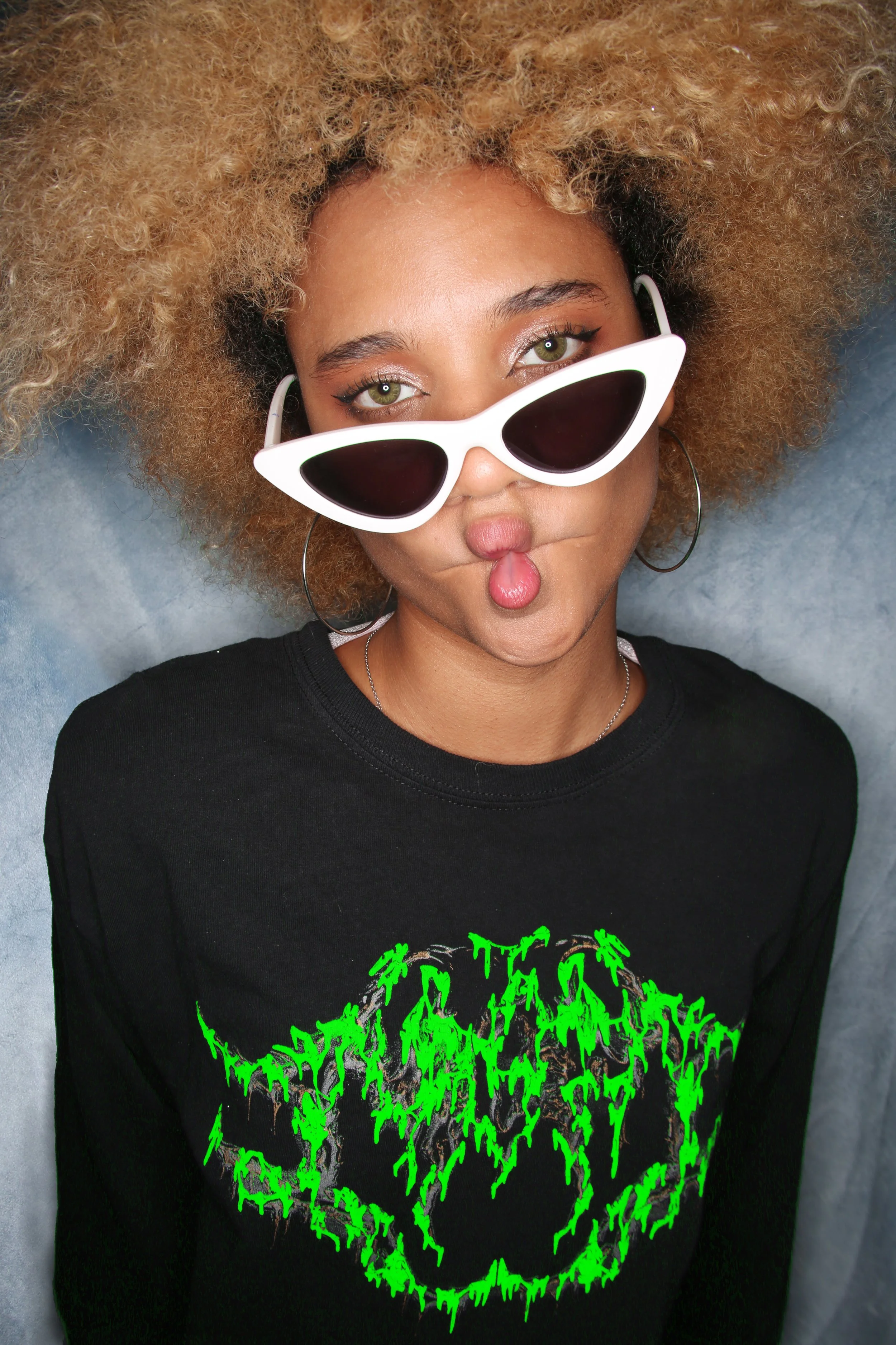 A woman with curly blonde hair wearing white sunglasses with black lenses, silver hoop earrings, a black shirt, and green text with a skull graphic on it.