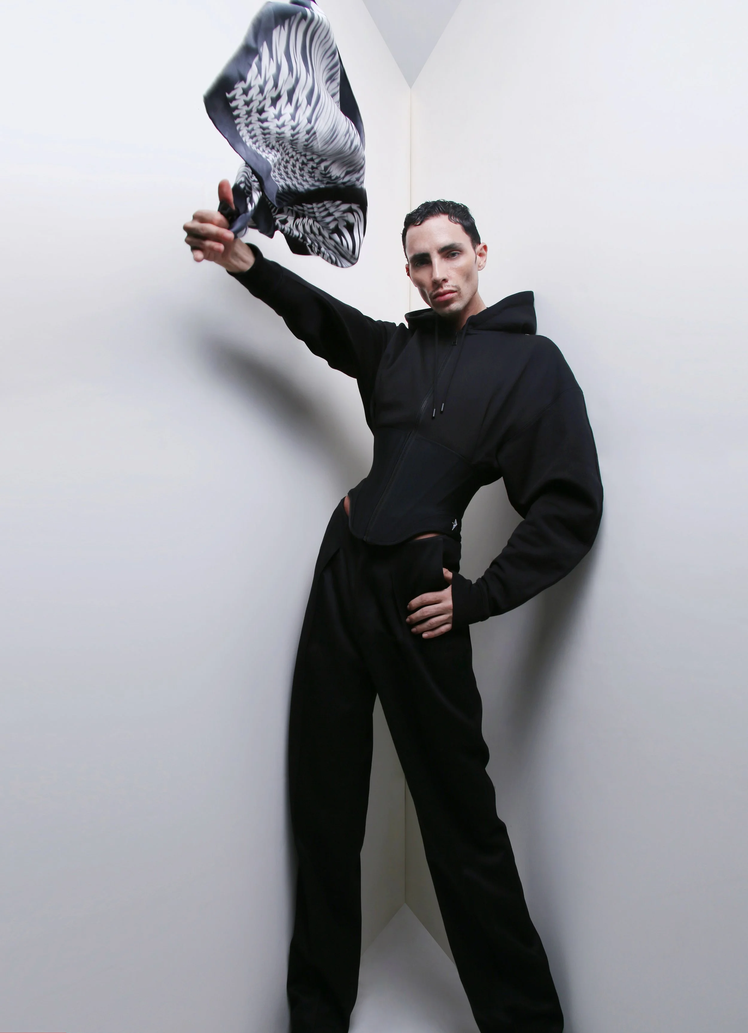A man with short dark hair wearing a black zip-up hoodie and black pants. He stands against white corner walls, striking a confident pose with one hand on his hip and the other holding a patterned fabric above his head.