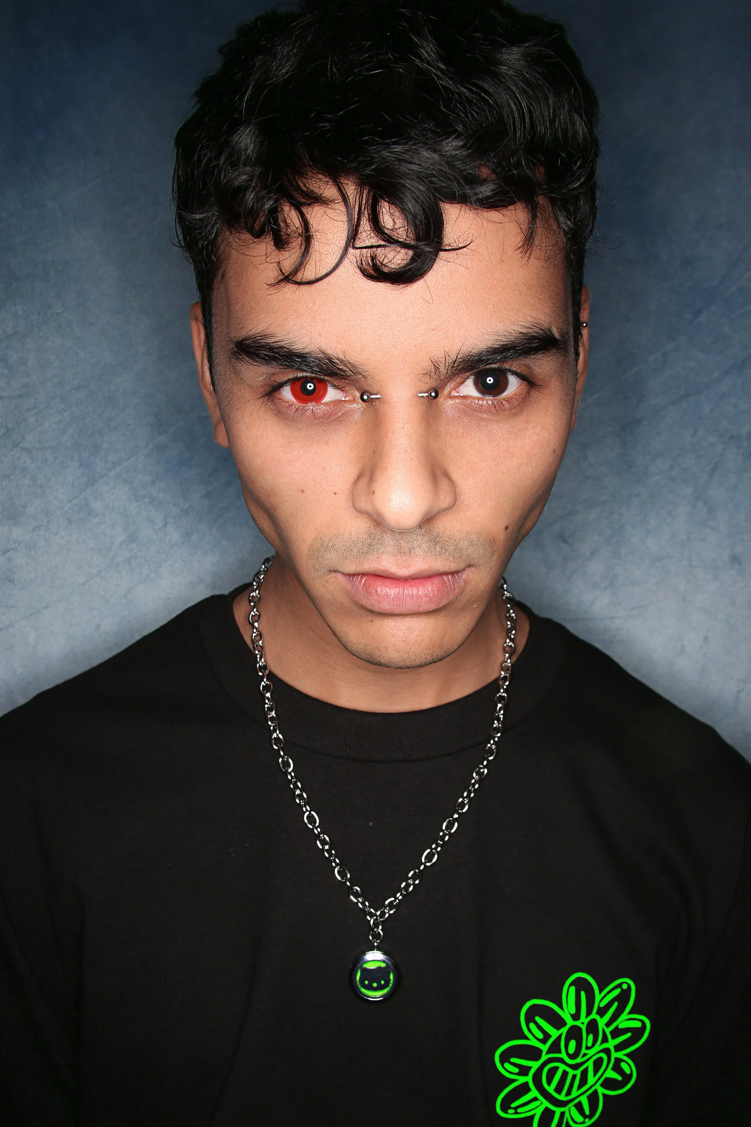 A young man with dark, curly hair and a serious expression, wearing a black shirt with a neon green smiley face necklace and a neon green doodle design on his shirt, against a dark background.