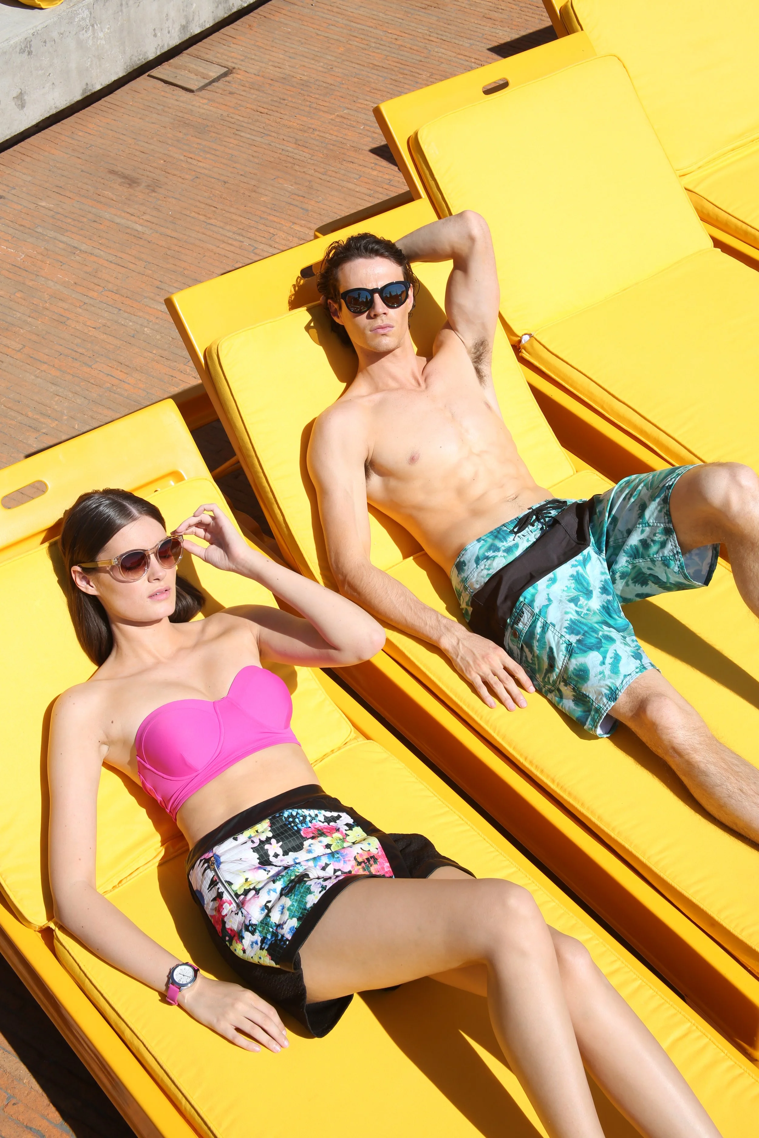 A man and a woman relaxing on yellow lounge chairs outdoors in sunlight. The man is shirtless, wearing sunglasses and patterned swim trunks. The woman is wearing sunglasses, a pink bikini top, and floral shorts.