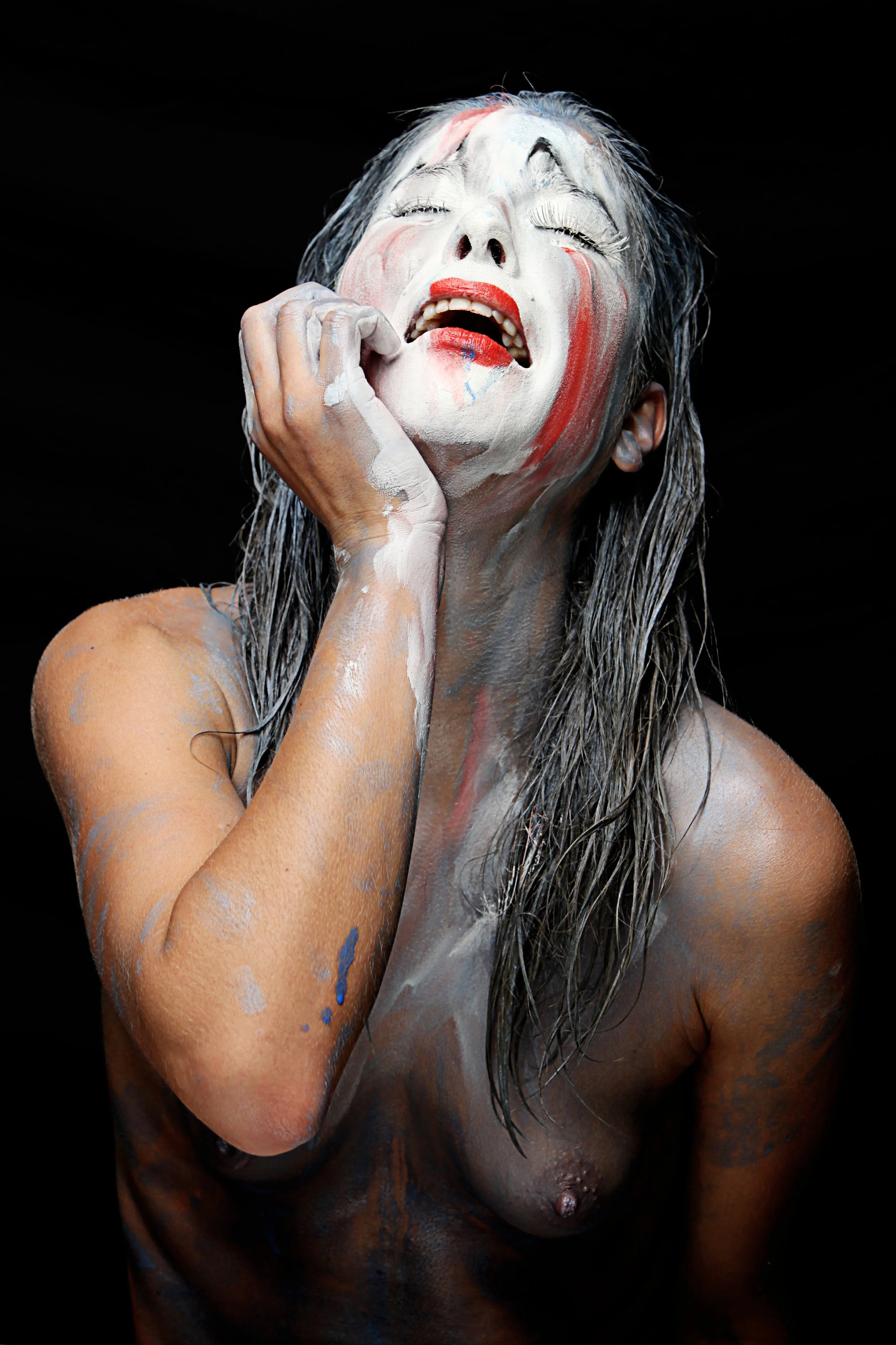 A woman with long wet hair and painted face, showing an expression of pain or anguish, against a black background.