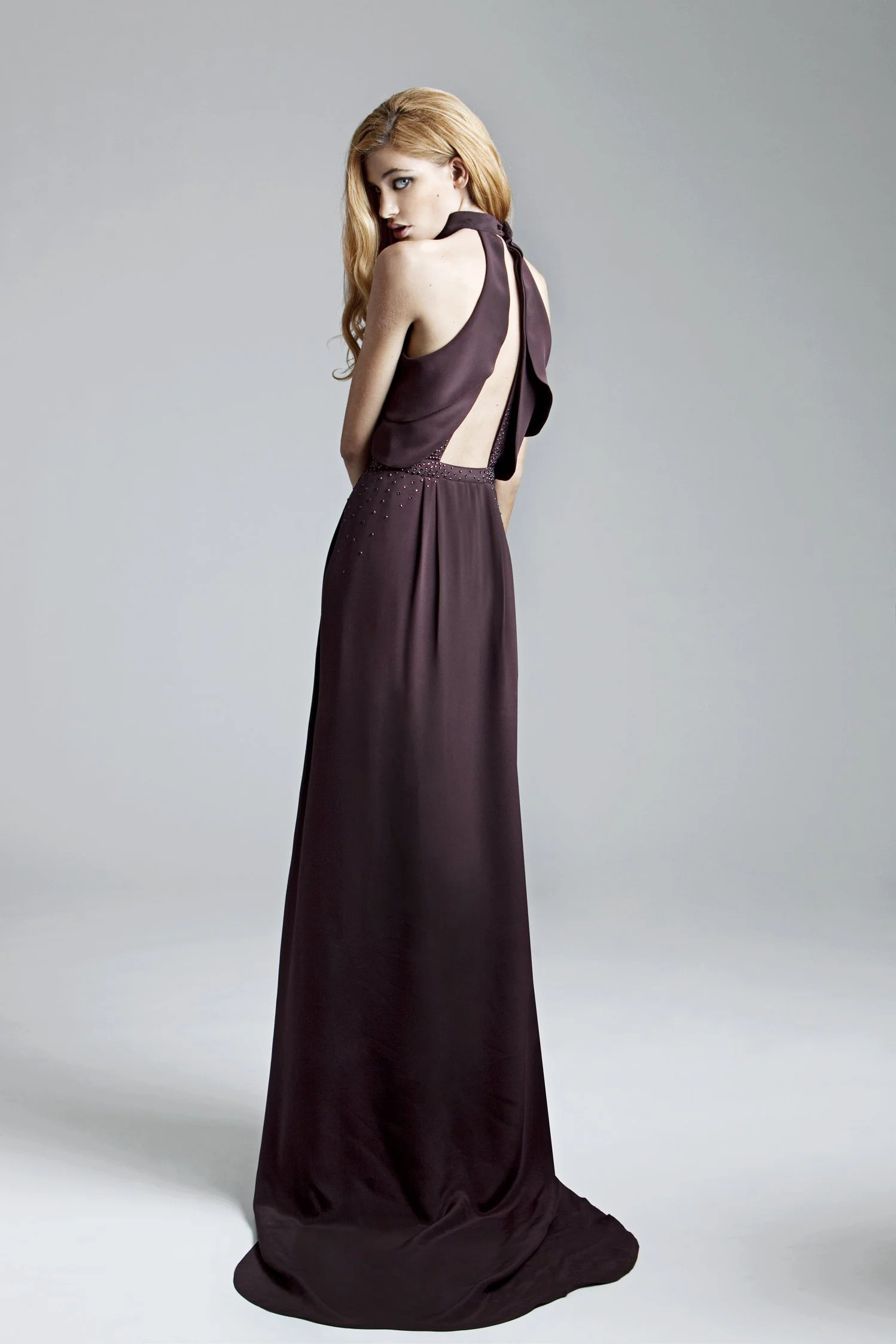 A woman with long red hair wearing a dark purple evening gown with an open back and tied bow at the neck, standing against a plain light gray background.