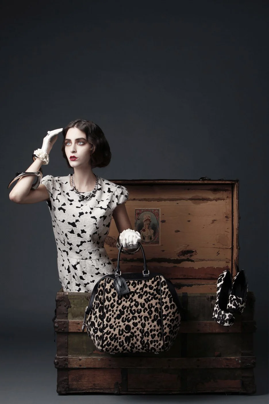 A woman with dark brown hair and fair skin wearing a black and white patterned dress, white gloves, and jewelry, stands next to a vintage wooden chest filled with designer leopard print bags and accessories, against a dark gray background.