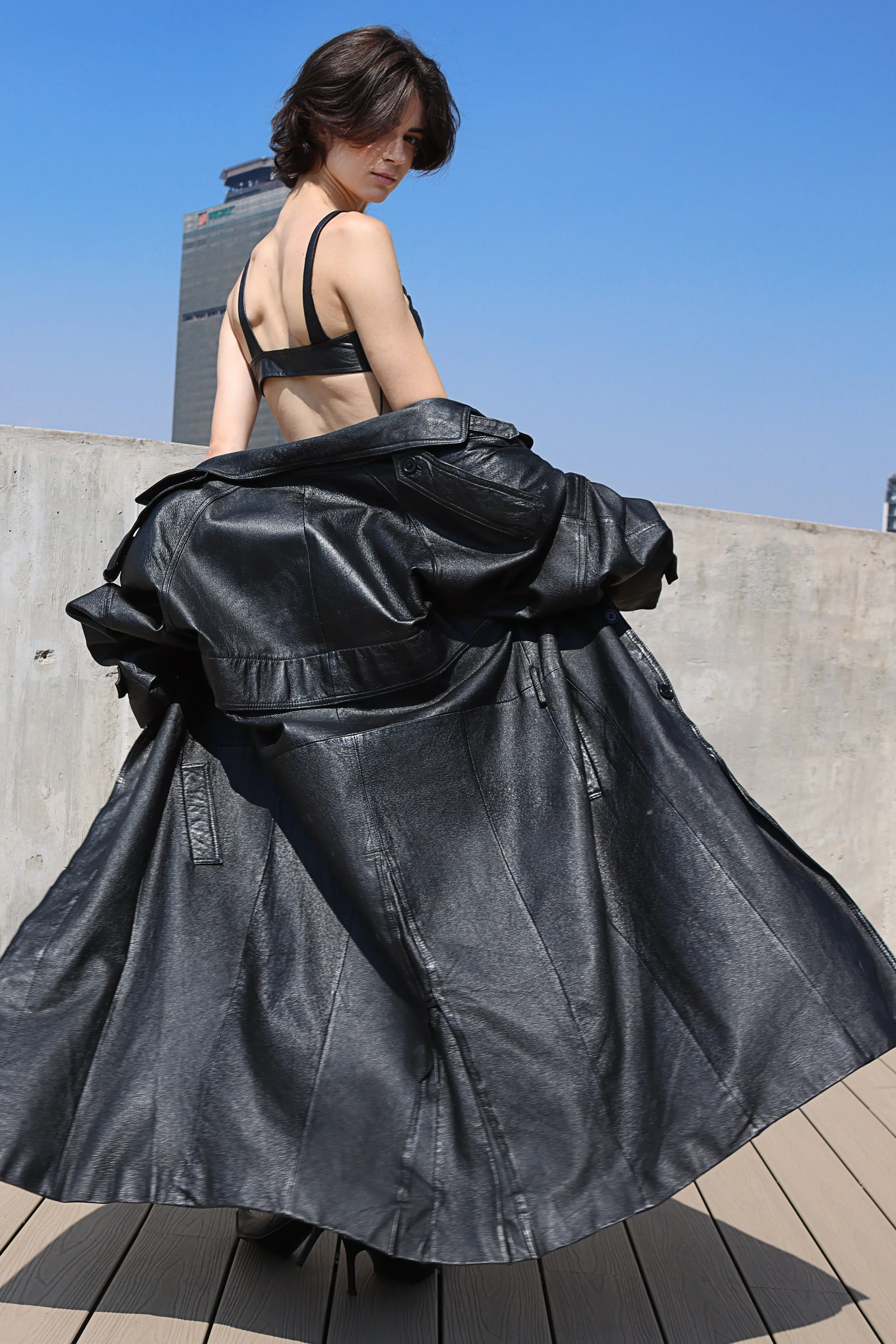 A woman with short brown hair in a black leather crop top and a long black leather skirt sitting on a concrete ledge against a clear blue sky, with a tall modern building in the background.