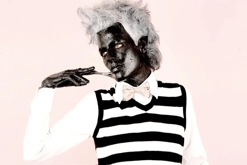 Person with dark skin tone, white hair, and black lipstick, wearing a black and white striped vest, white shirt, and a bow tie, posing with hand near face against a plain background.