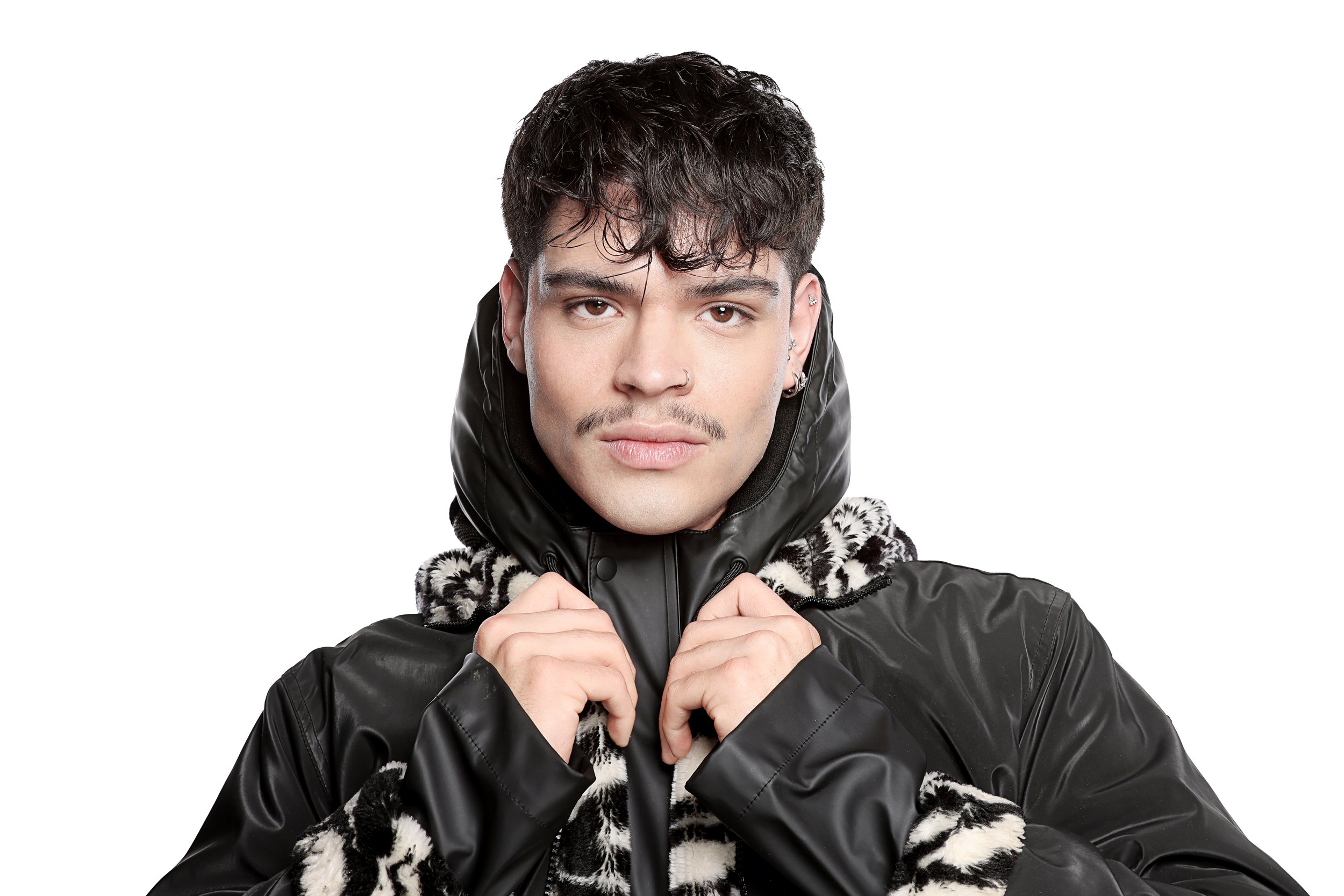 Young man with dark, curly hair and earrings, wearing a black jacket with a leopard print hoodie, holding the collar of his jacket with both hands, against a plain white background.